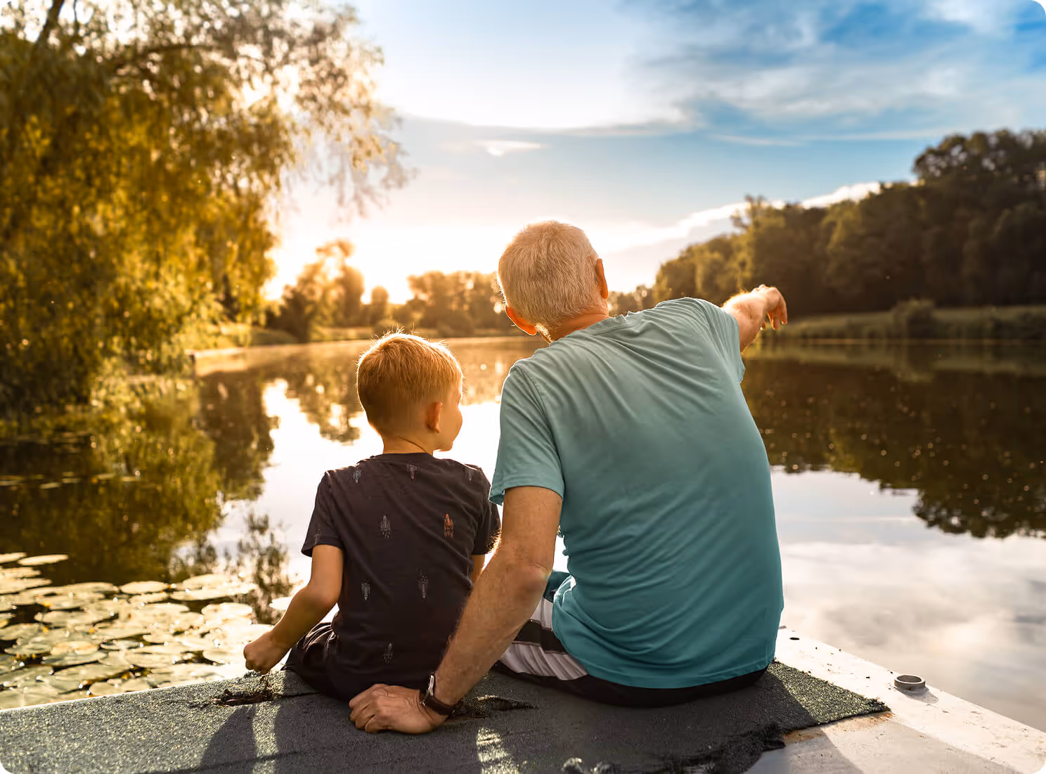 A small child and a grown man sitting next to a pond at sunset.