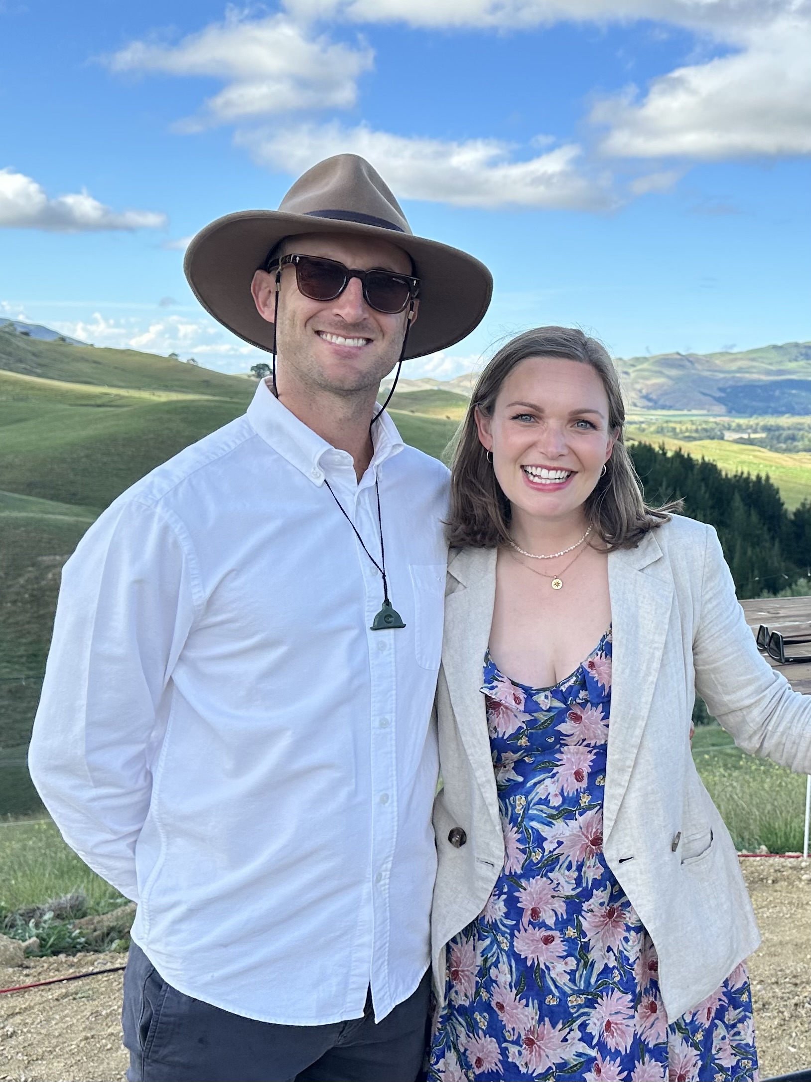 Smiling man in hat and sunglasses with woman in floral dress and blazer in green hilly landscape.