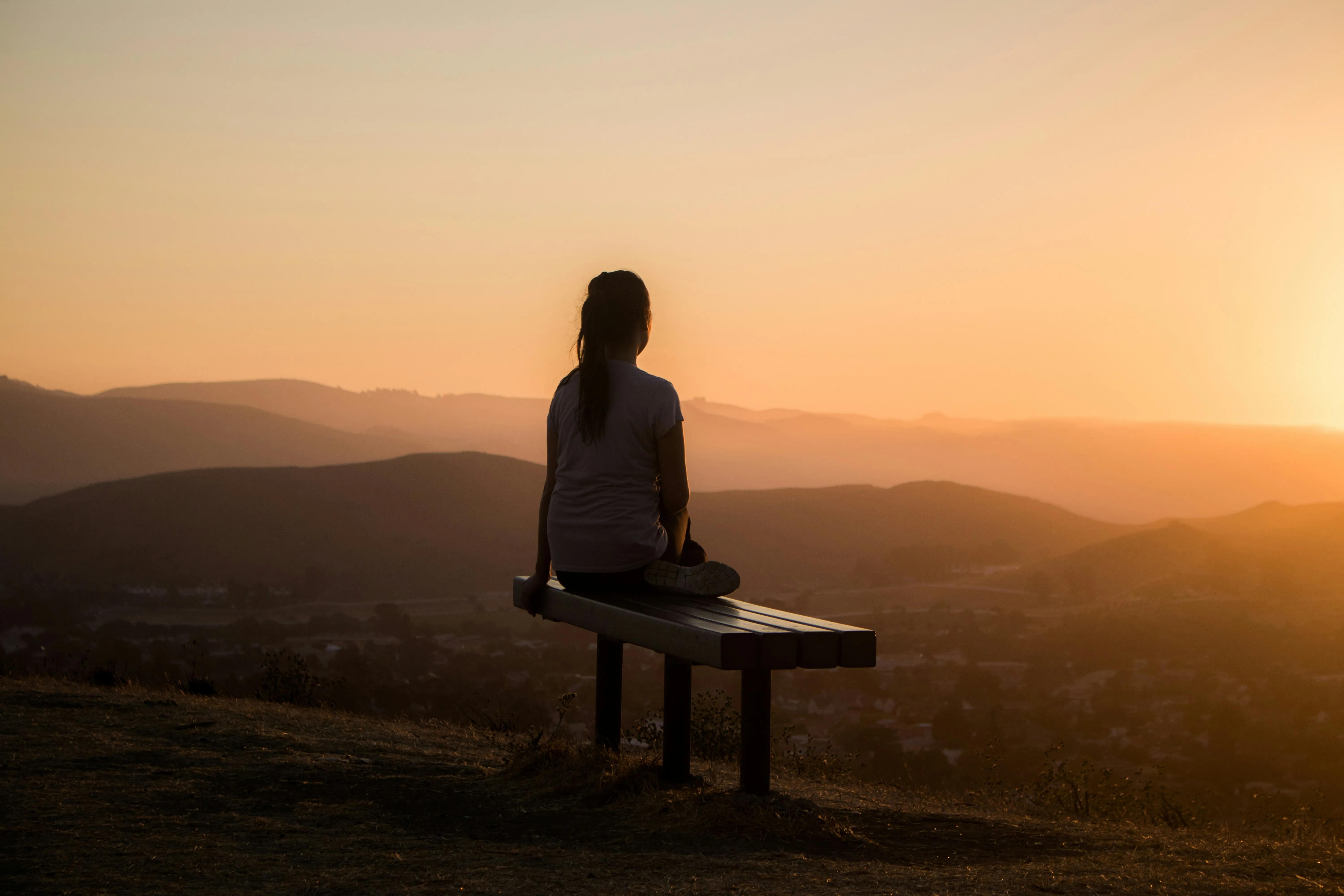 Person sitting on a bench overlooking hills and a sunset with an orange sky.