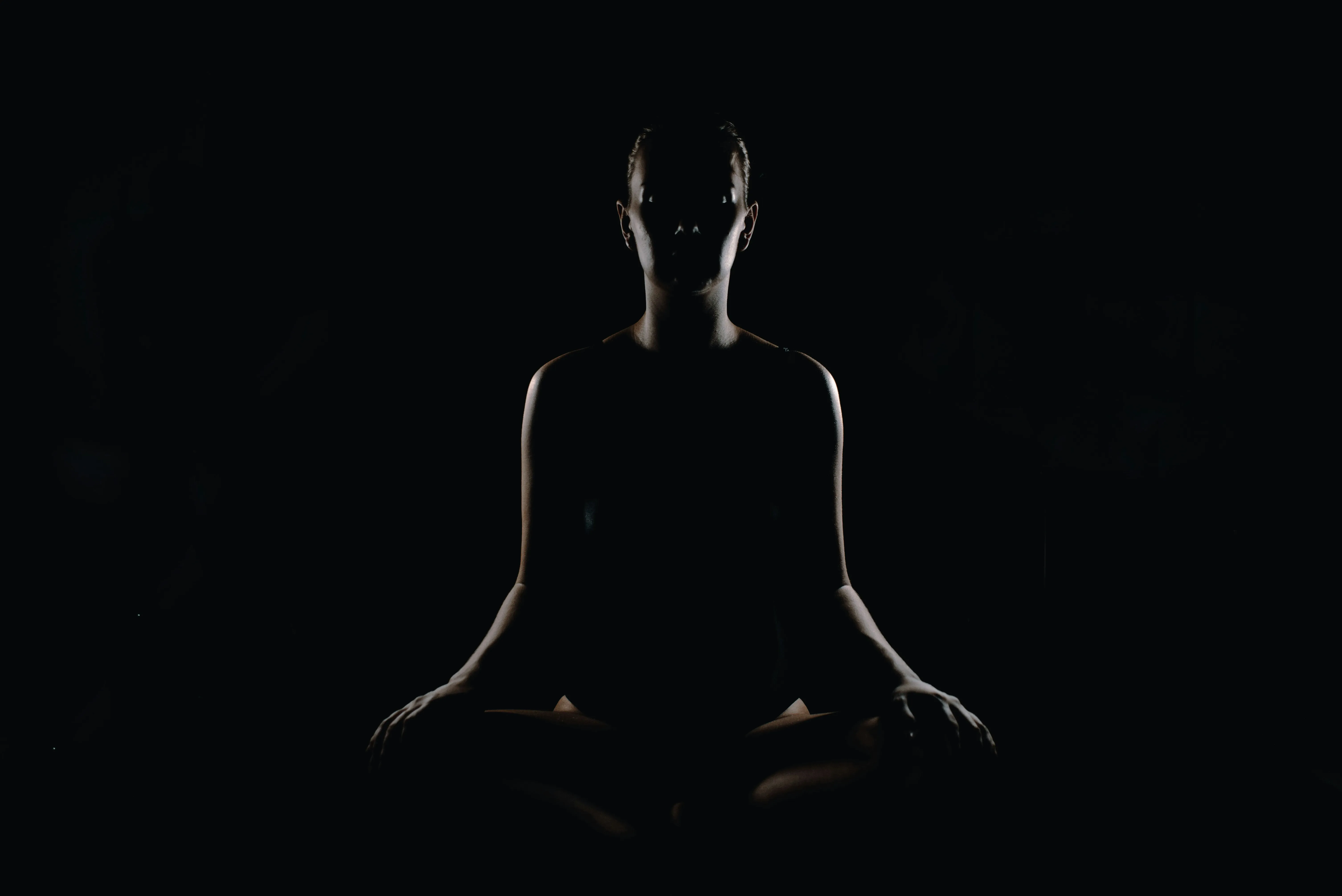 Silhouetted person seated cross-legged in meditation against a black background with subtle lighting.
