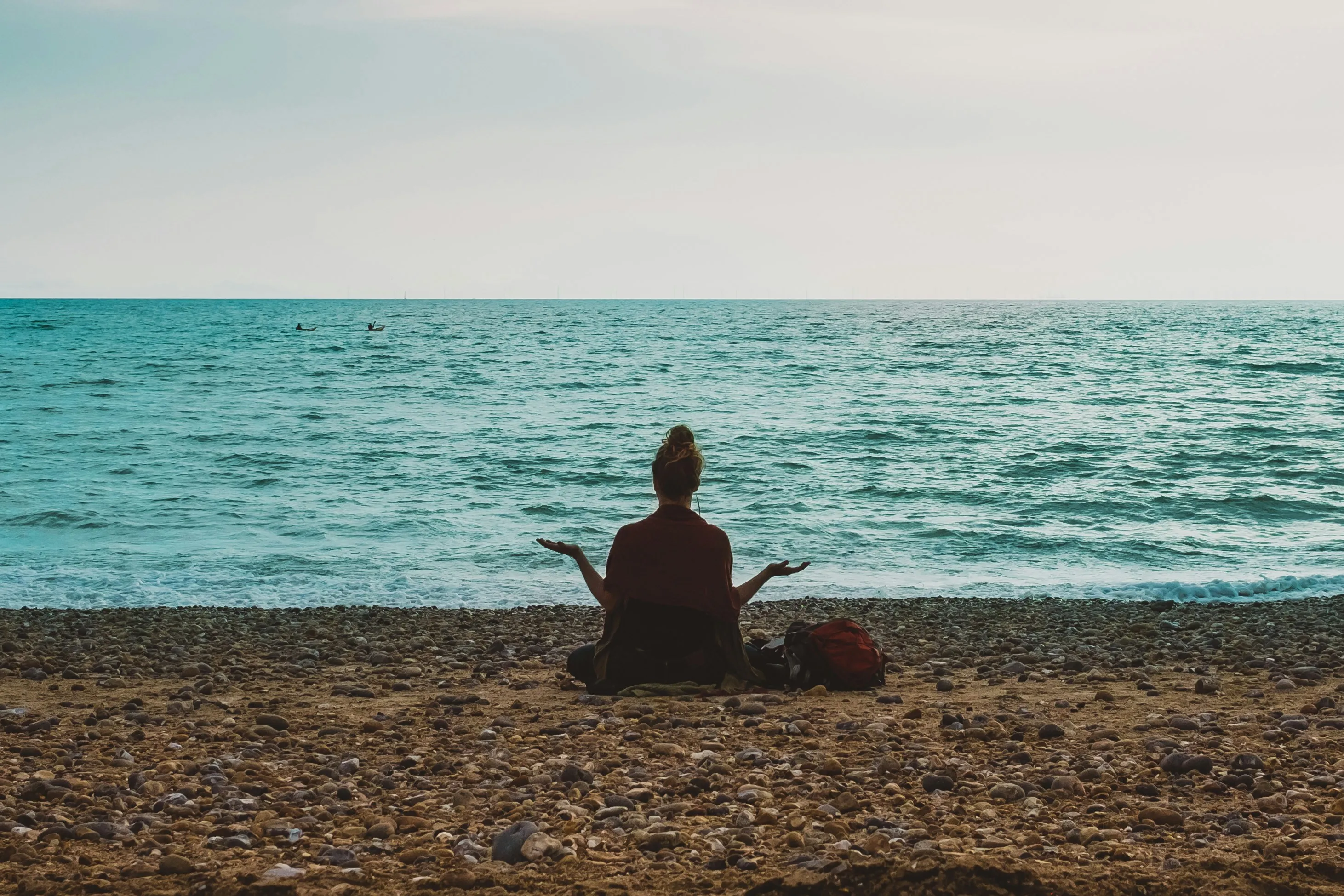 Person sitting cross-legged on a rocky beach facing the ocean with arms open wide.