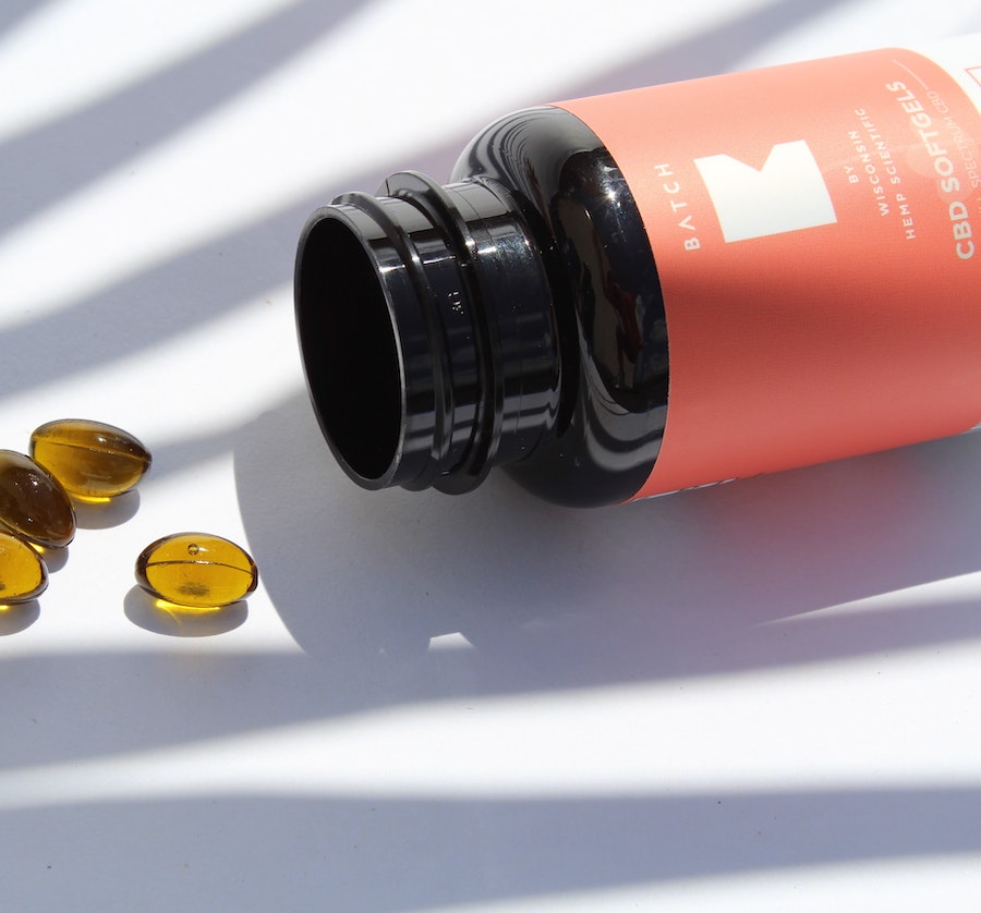 Black bottle with a pink label lying on its side with several amber CBD softgel capsules spilled on a white surface.