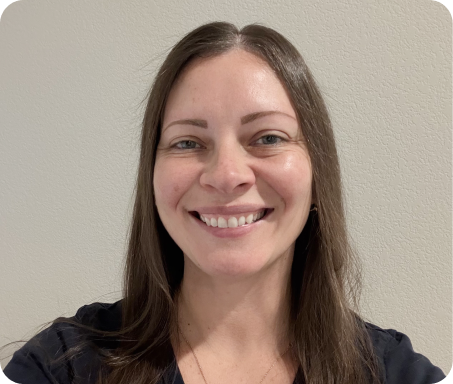Smiling woman with long brown hair wearing a black top against a light gray wall.