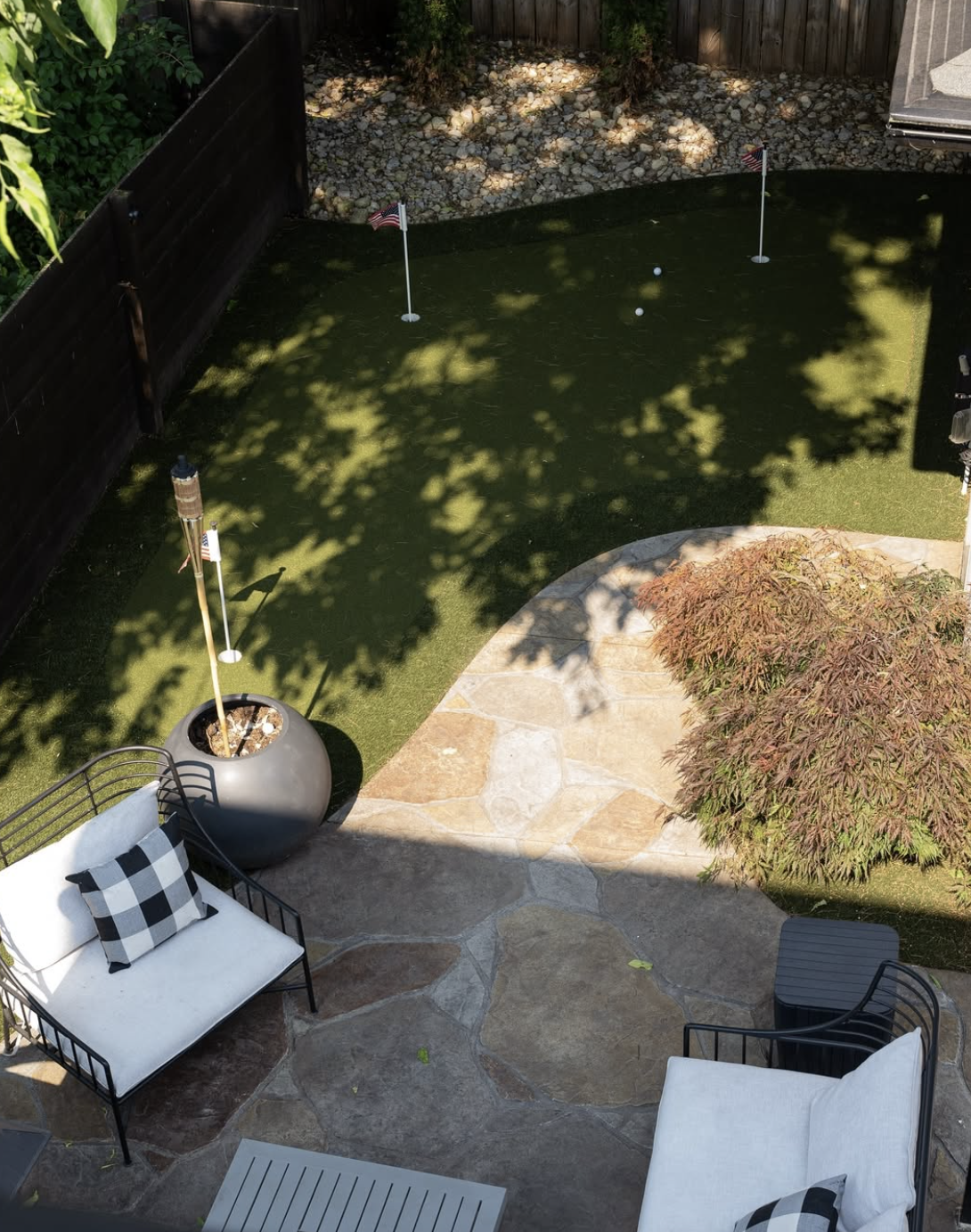Backyard patio with cushioned chairs, a putting green with three golf holes and balls, tiki torch, and a small tree casting shadows.
