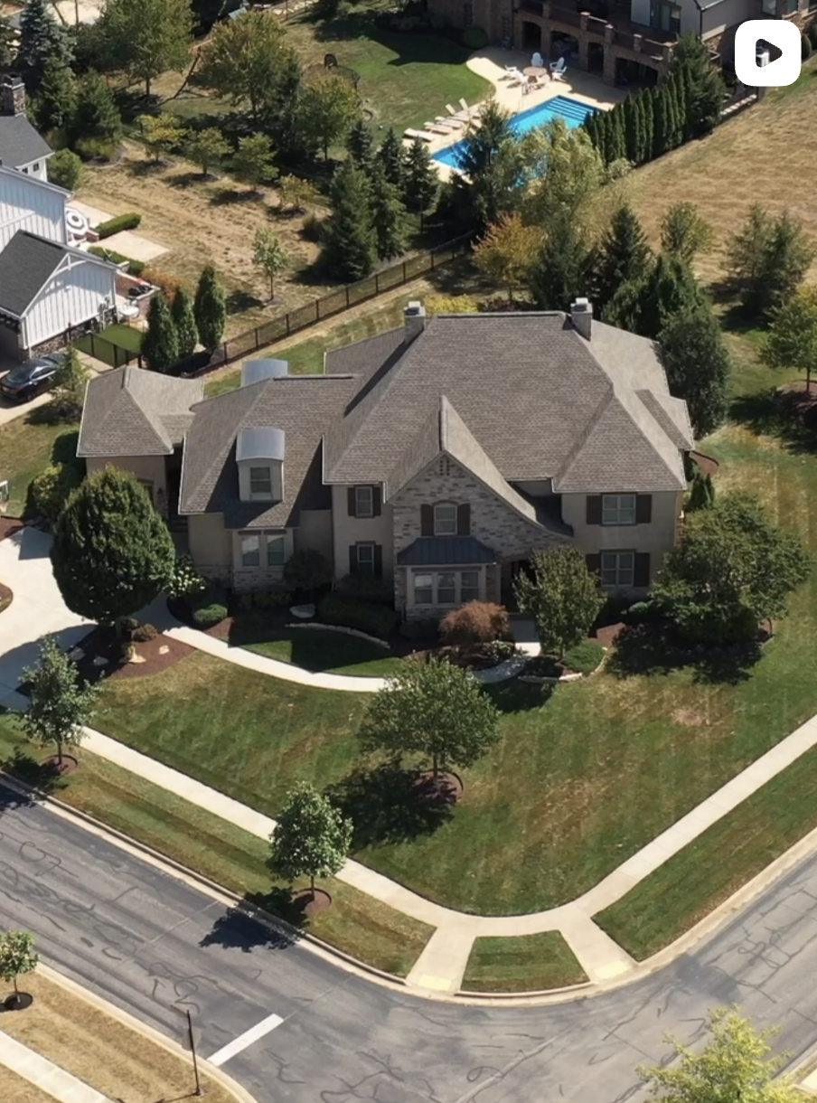 Aerial view of a large two-story suburban house on a corner lot with manicured lawn, surrounding trees, and a fenced backyard with a swimming pool.