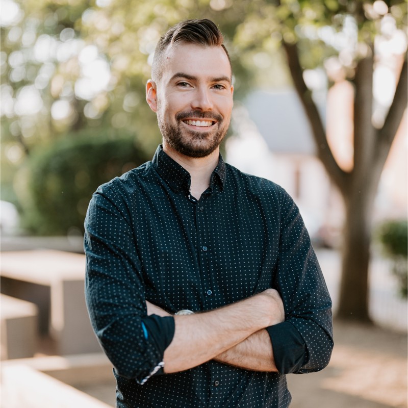 Smiling man with beard and short hair wearing a black patterned shirt, standing outdoors with arms crossed.