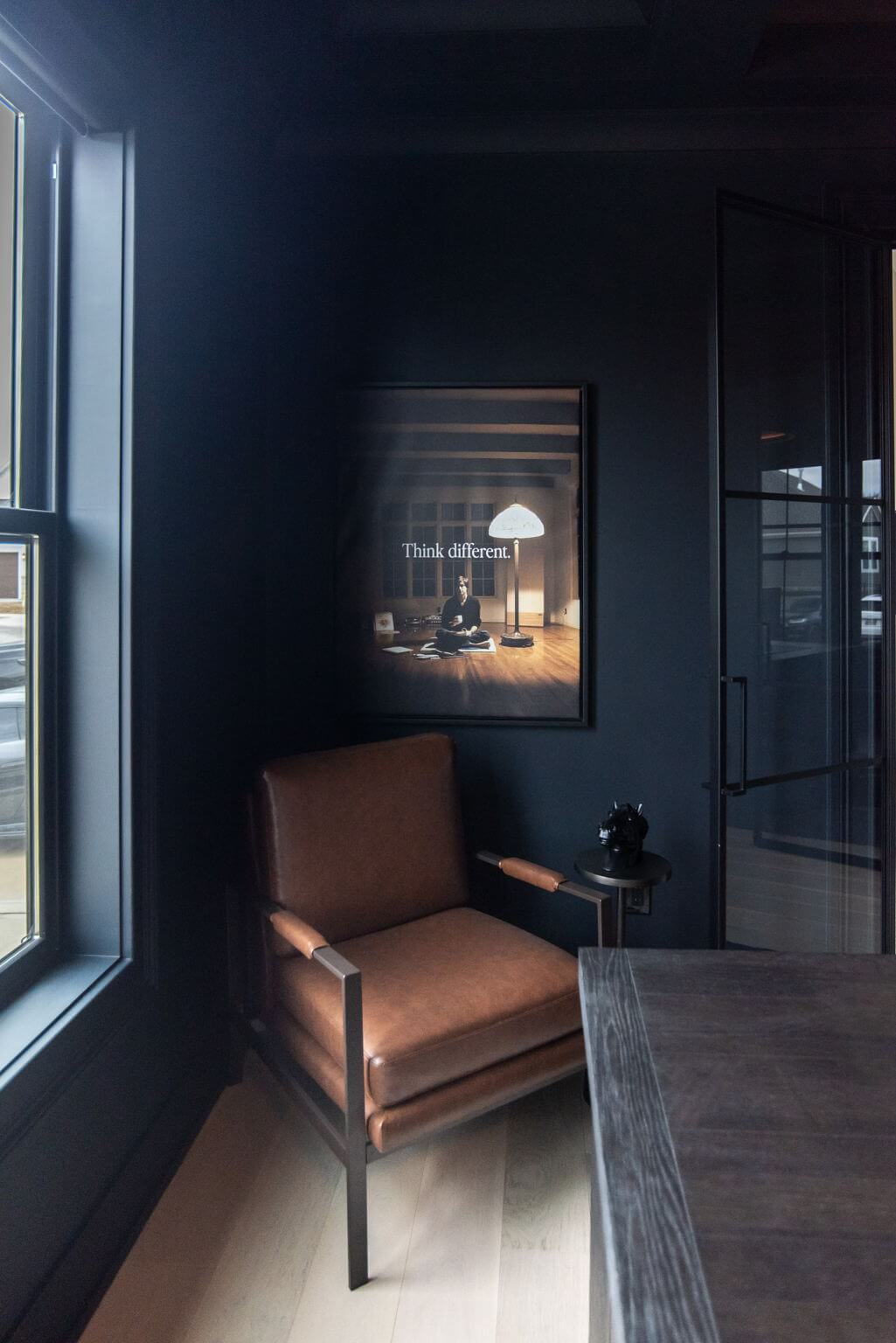 Brown leather chair next to a window in a room with dark blue walls and a framed 'Think different.' poster.