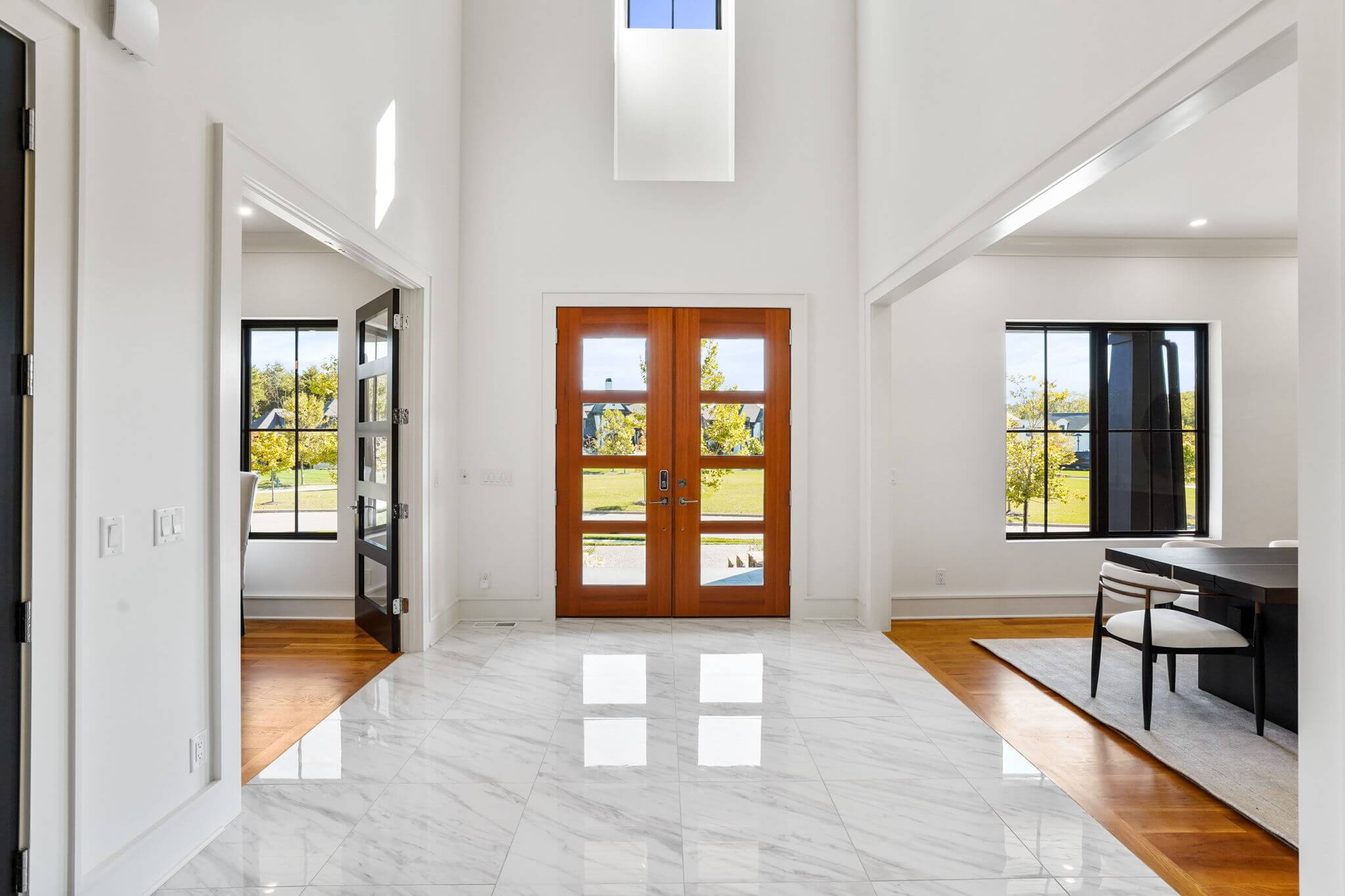Bright modern entryway with polished white marble floor, wooden double front doors, and adjacent rooms with large windows.