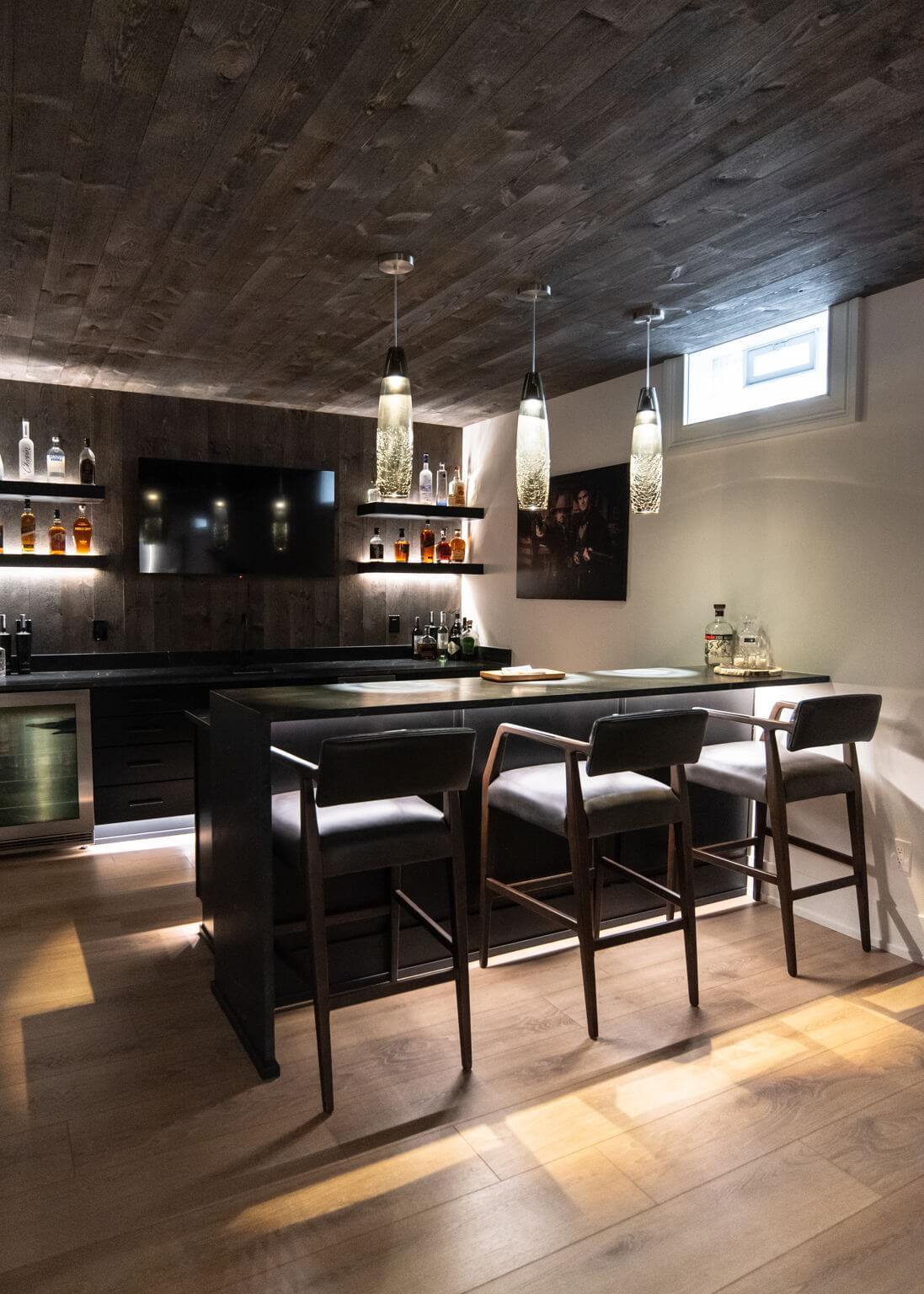 Modern home bar with three wooden bar stools, pendant lights, shelves with liquor bottles, and a wall-mounted TV.