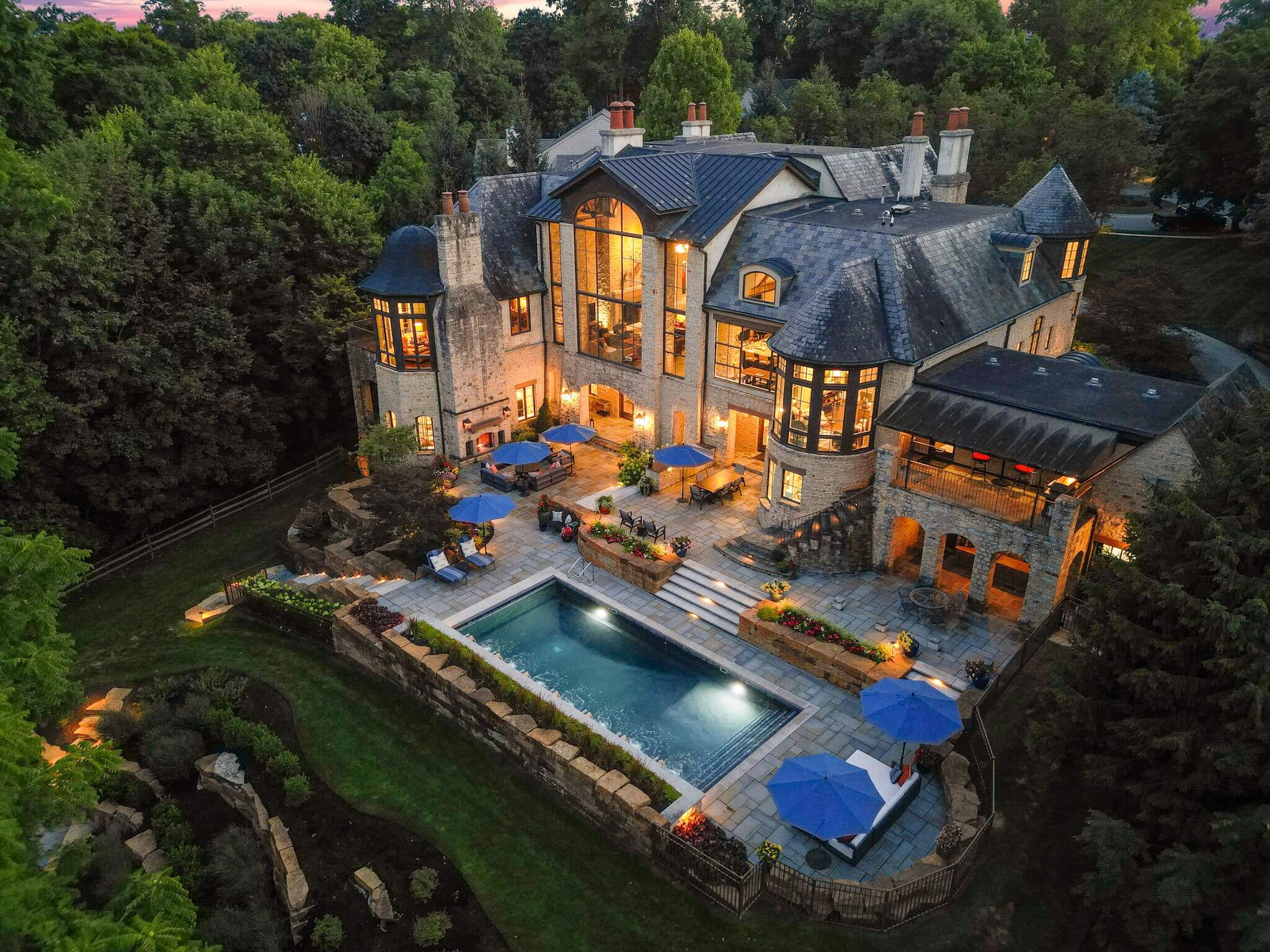A large stone mansion with multiple chimneys and tall windows overlooking a lit swimming pool surrounded by a patio with blue umbrellas and lounge chairs in a wooded area at dusk.