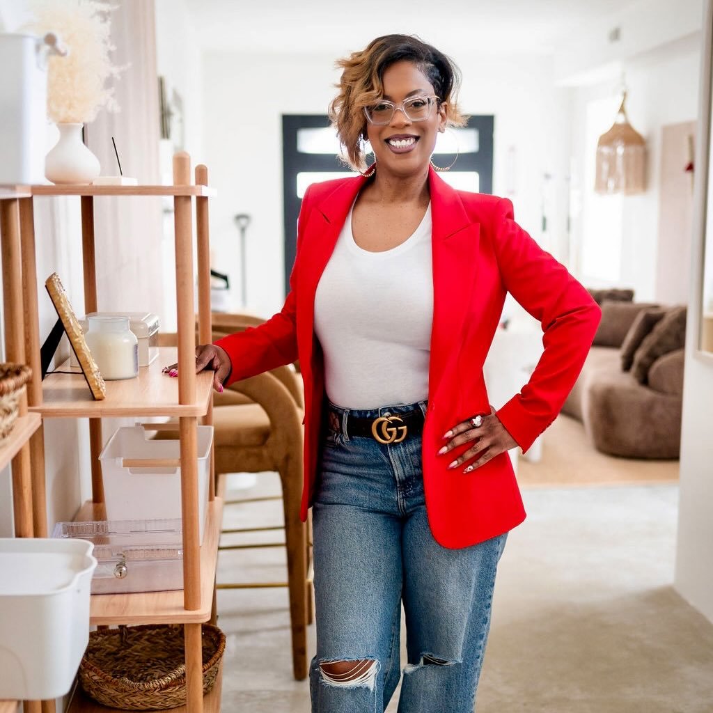 Smiling woman wearing glasses, a red blazer, white top, and ripped jeans, standing indoors with one hand on a wooden shelf and the other on her hip.