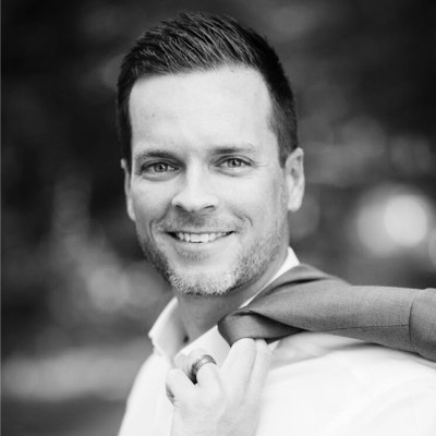 Smiling man with short hair holding a jacket over his shoulder in a blurred outdoor setting.
