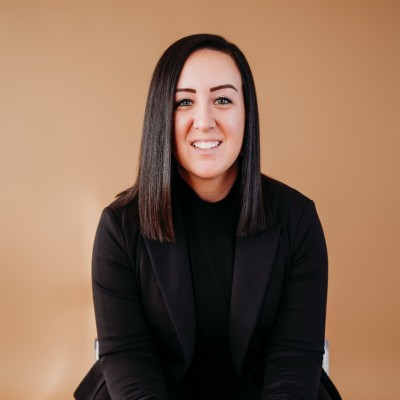 Smiling woman with straight dark hair wearing a black blazer and top against a tan background.
