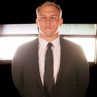 Serious man wearing a black suit and tie standing in front of bright rectangular lights against a black background.