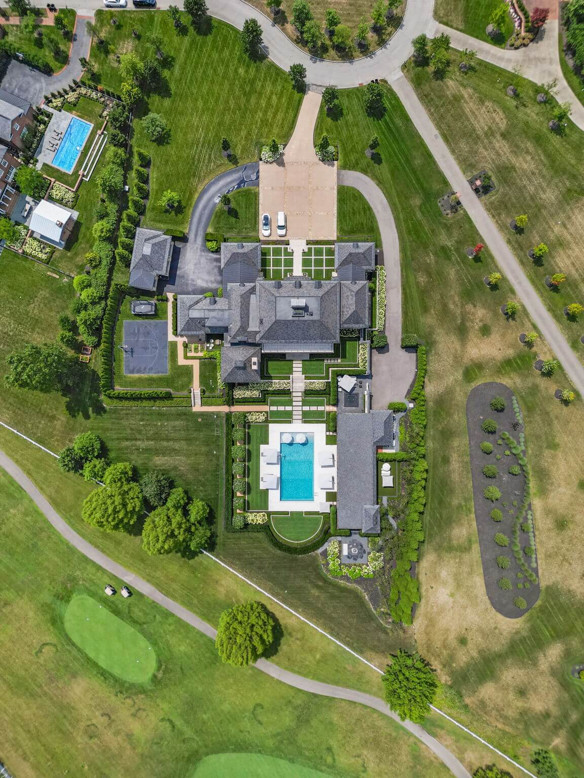 Aerial view of a large estate featuring a main house, a swimming pool, tennis court, and a nearby small pool surrounded by green lawns and trees.