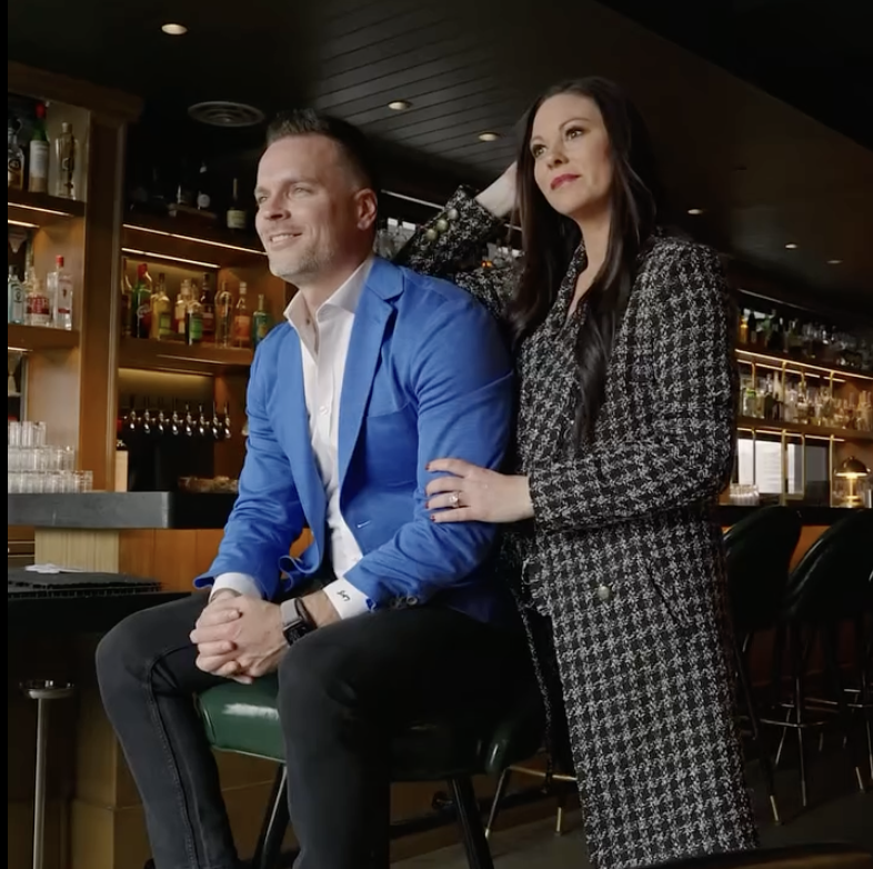 Man in blue jacket sitting on a bar stool and smiling, woman in patterned coat standing beside him with her hand on his shoulder inside a bar.
