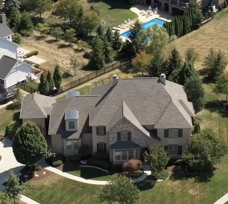 Aerial view of a large suburban house with a well-kept lawn, surrounded by trees and neighboring homes, with a swimming pool visible in the background.
