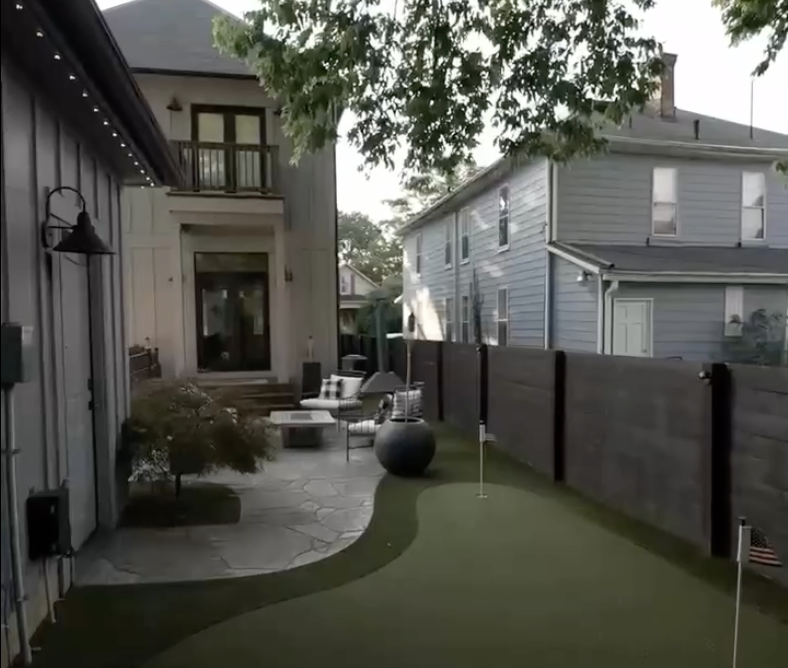 Backyard with a putting green, stone patio, outdoor seating area, and adjacent buildings with wooden fences.