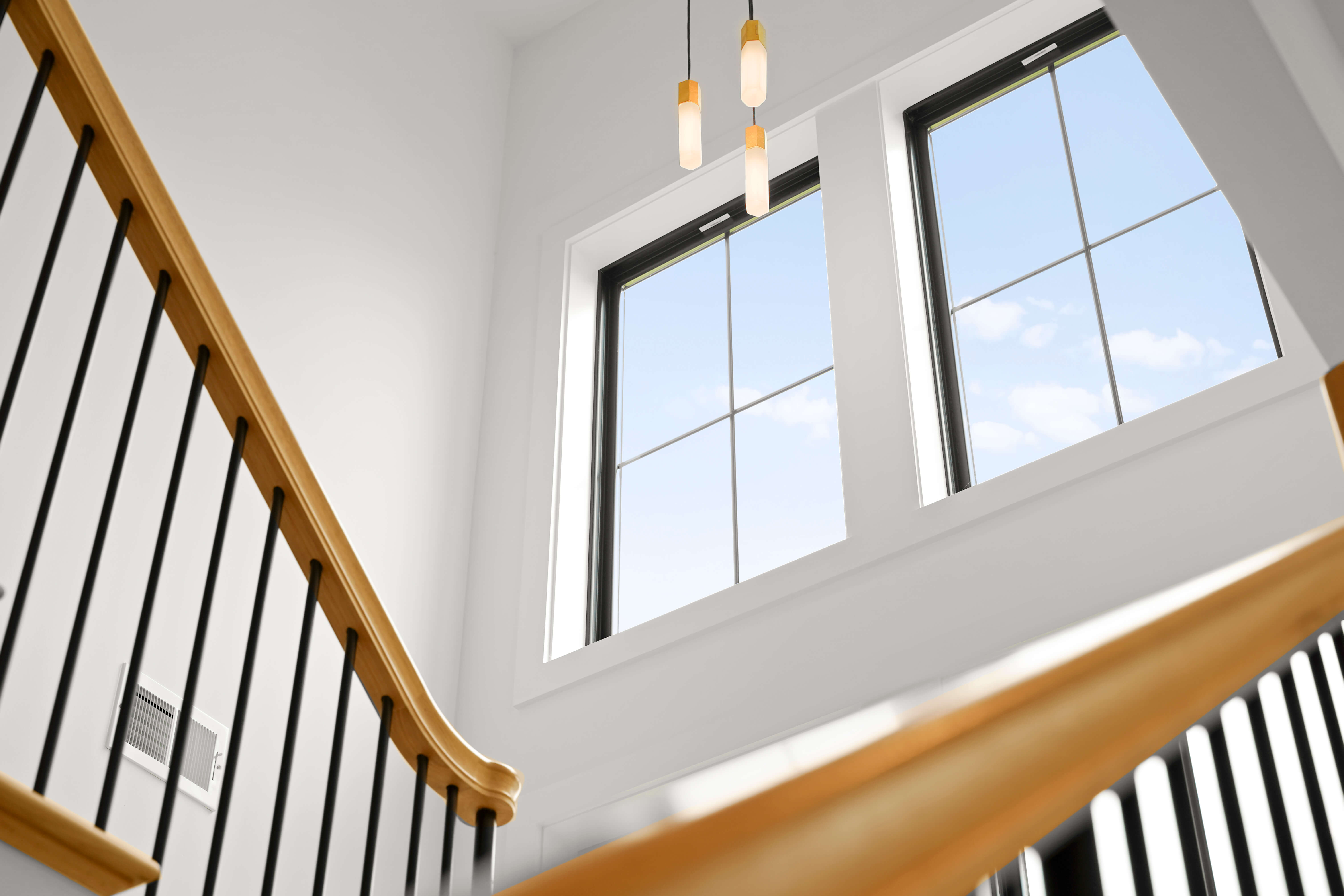 Interior stairway with wooden handrails, black metal balusters, large windows showing blue sky and clouds, and three hanging pendant lights.
