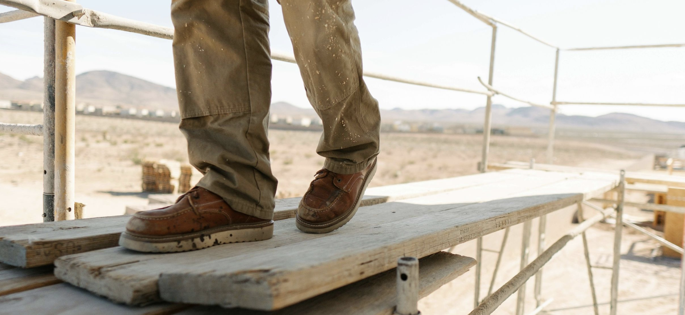 Person wearing brown shoes and khaki pants walking on wooden planks on a construction scaffold in a desert area.