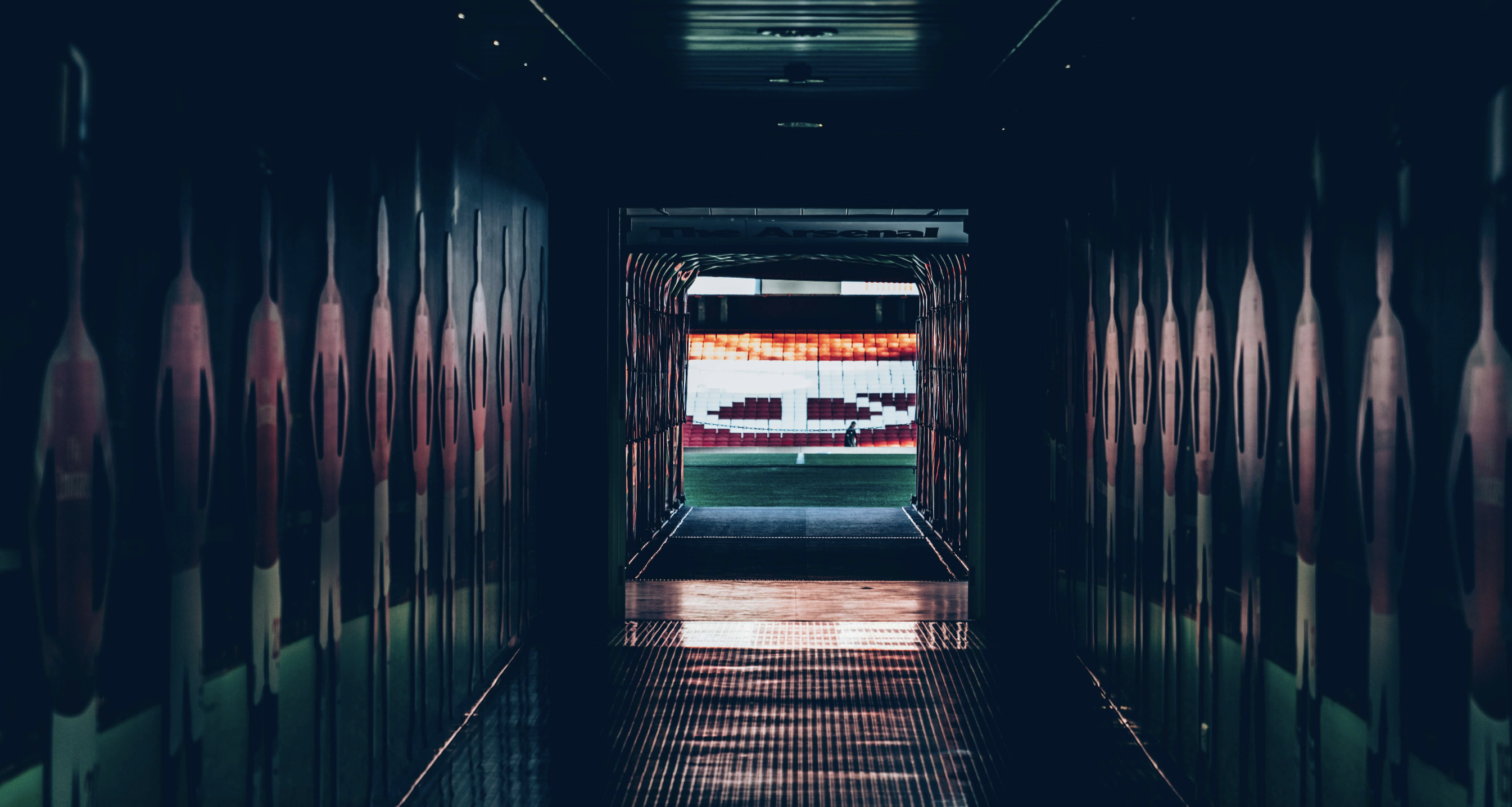 A dark tunnel lined with player silhouettes leads to a brightly lit football pitch and stands.