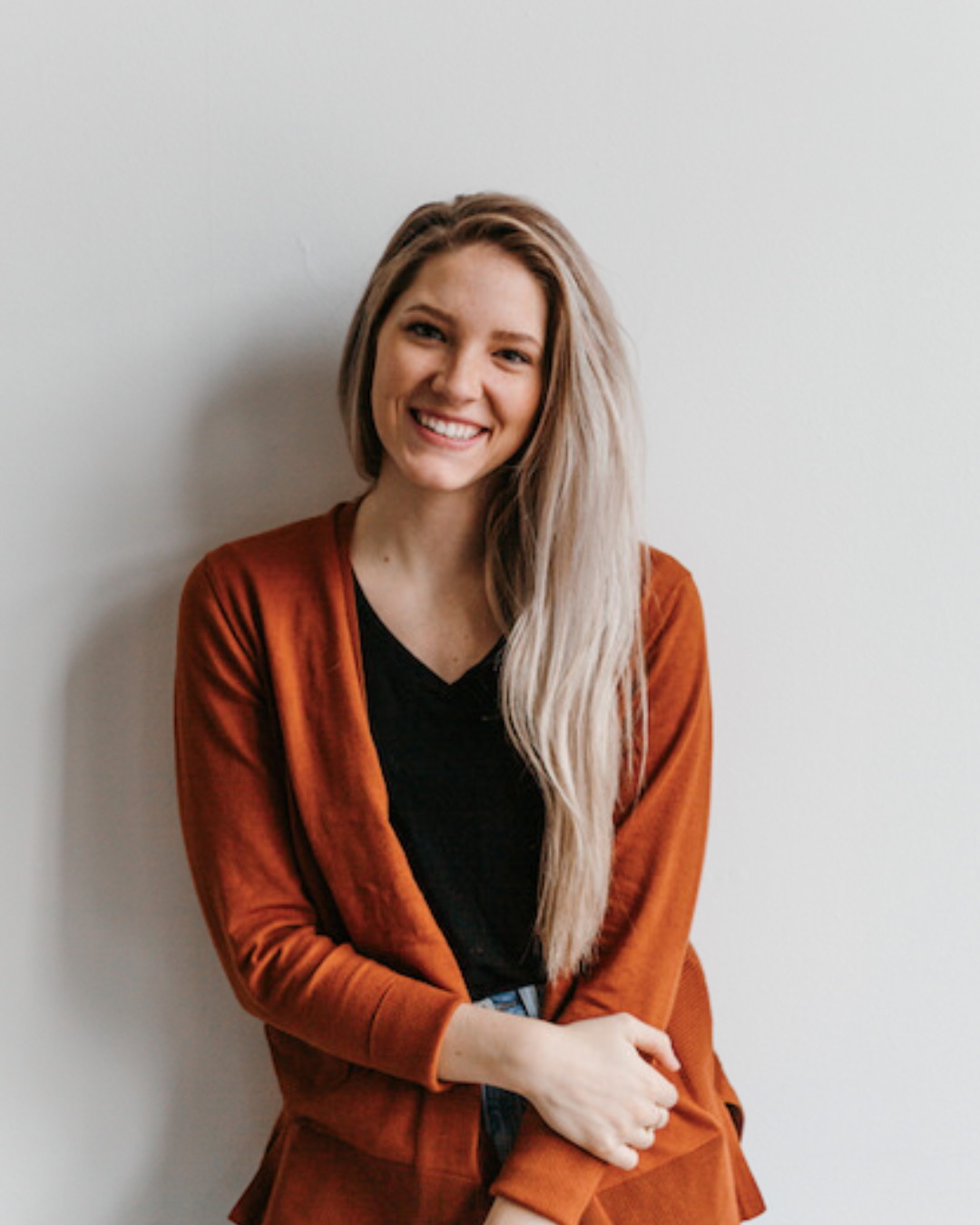 Smiling young woman with long blonde hair wearing a rust-colored cardigan and black shirt, standing against a white wall.