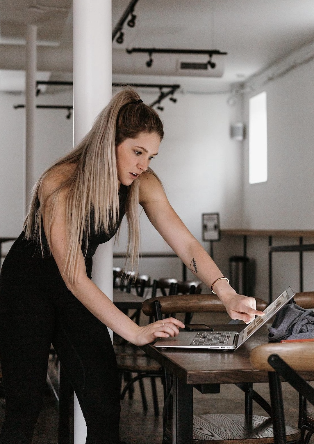 Woman with long blond hair tied in a ponytail leaning over a wooden table and interacting with a laptop.