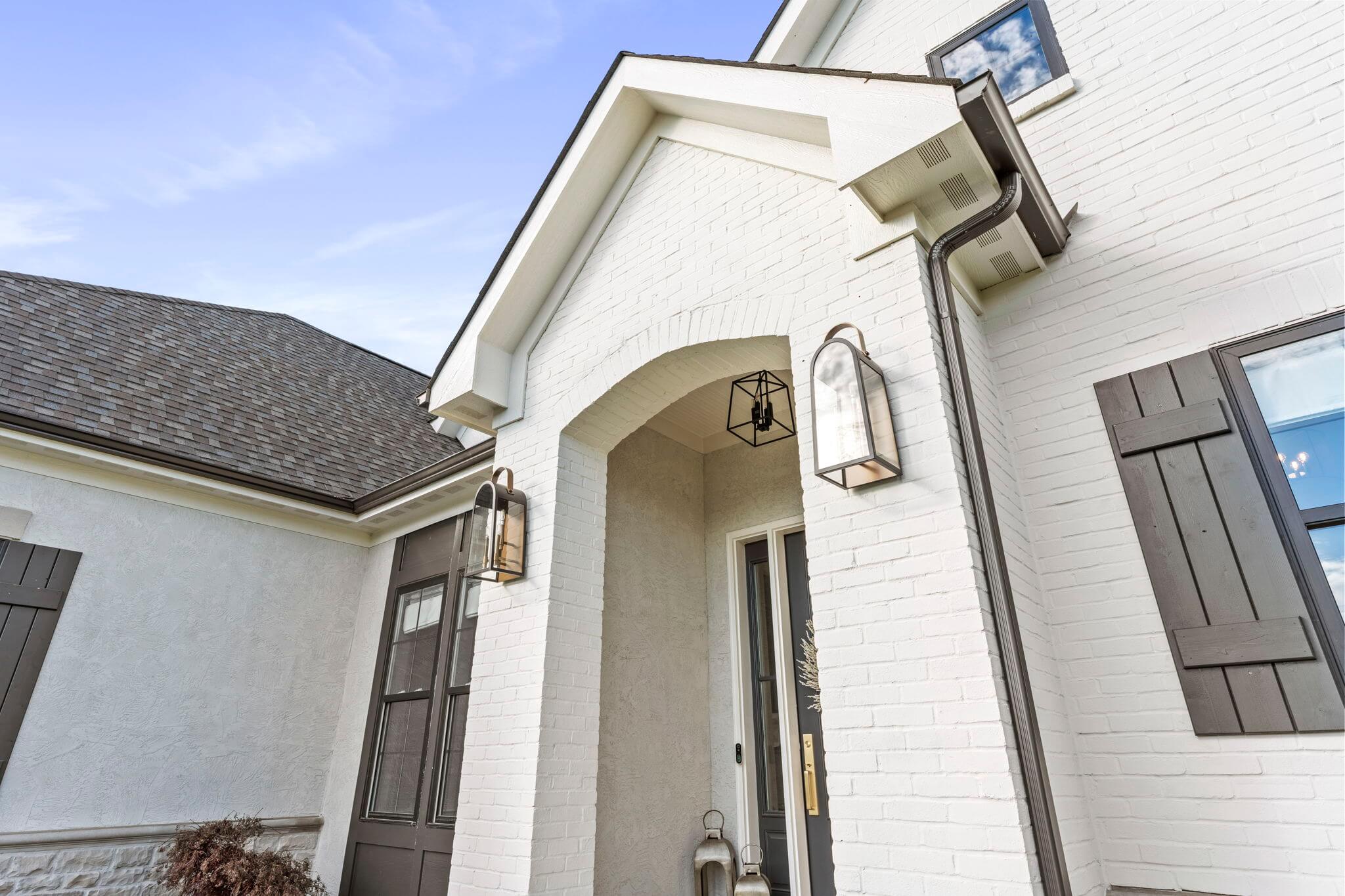 White brick house entryway with arched porch, black shutters, and modern wall lanterns on either side of the door.