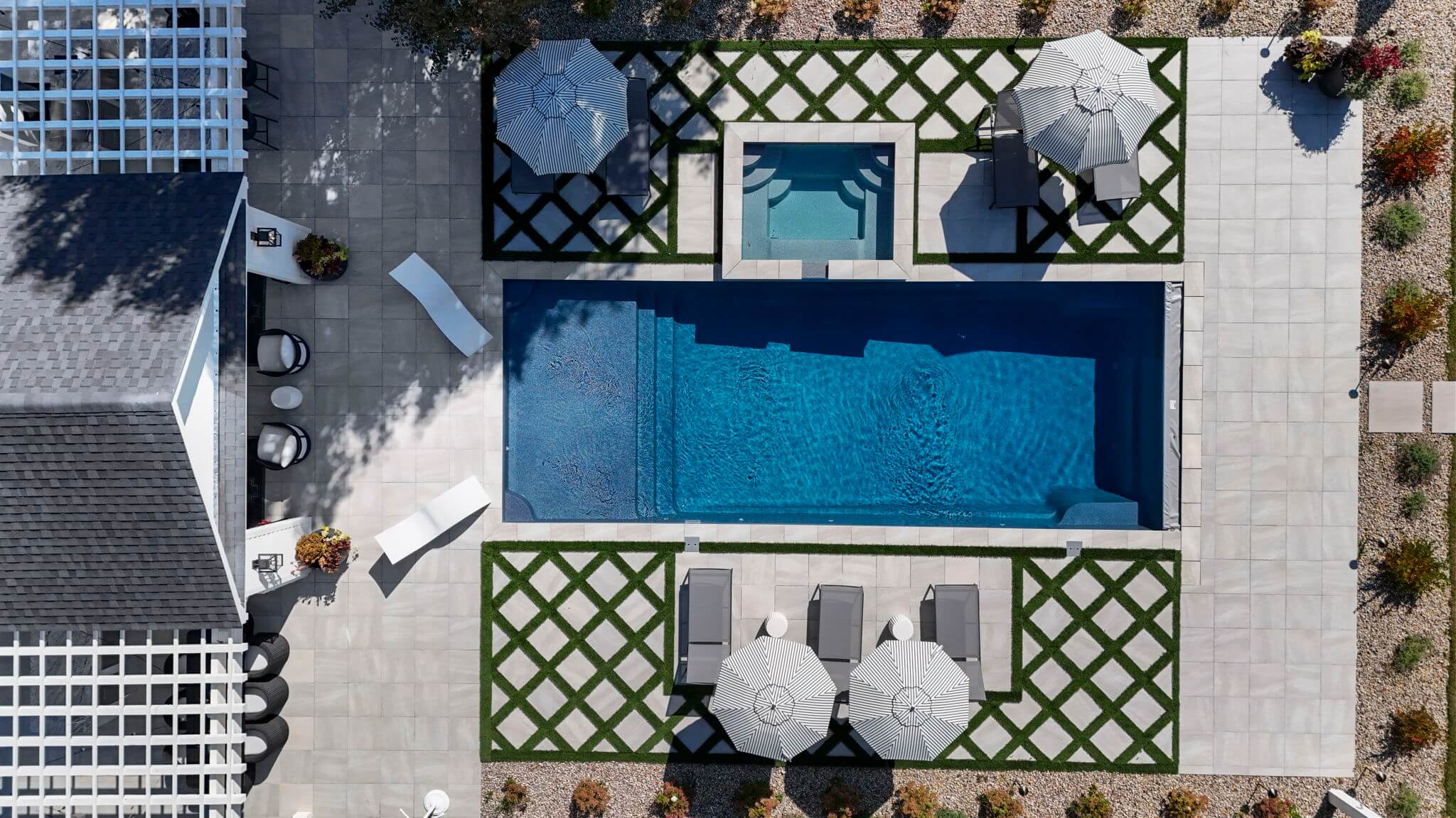 Aerial view of a modern backyard with a rectangular swimming pool, adjacent hot tub, lounge chairs under striped umbrellas, and a patterned stone patio with greenery.