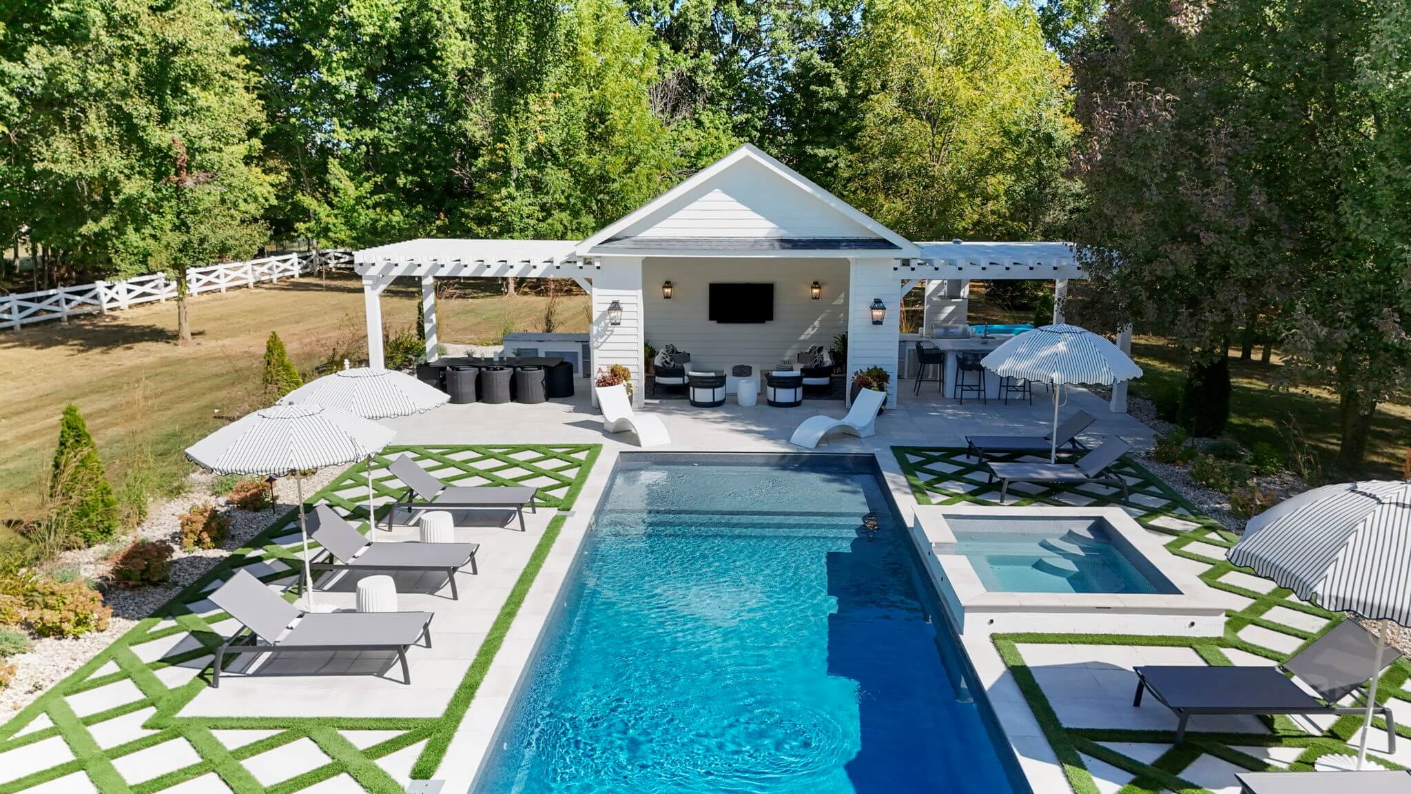 Modern backyard pool area with blue rectangular pool, adjacent hot tub, lounge chairs with umbrellas, and a white pergola with seating and outdoor kitchen.