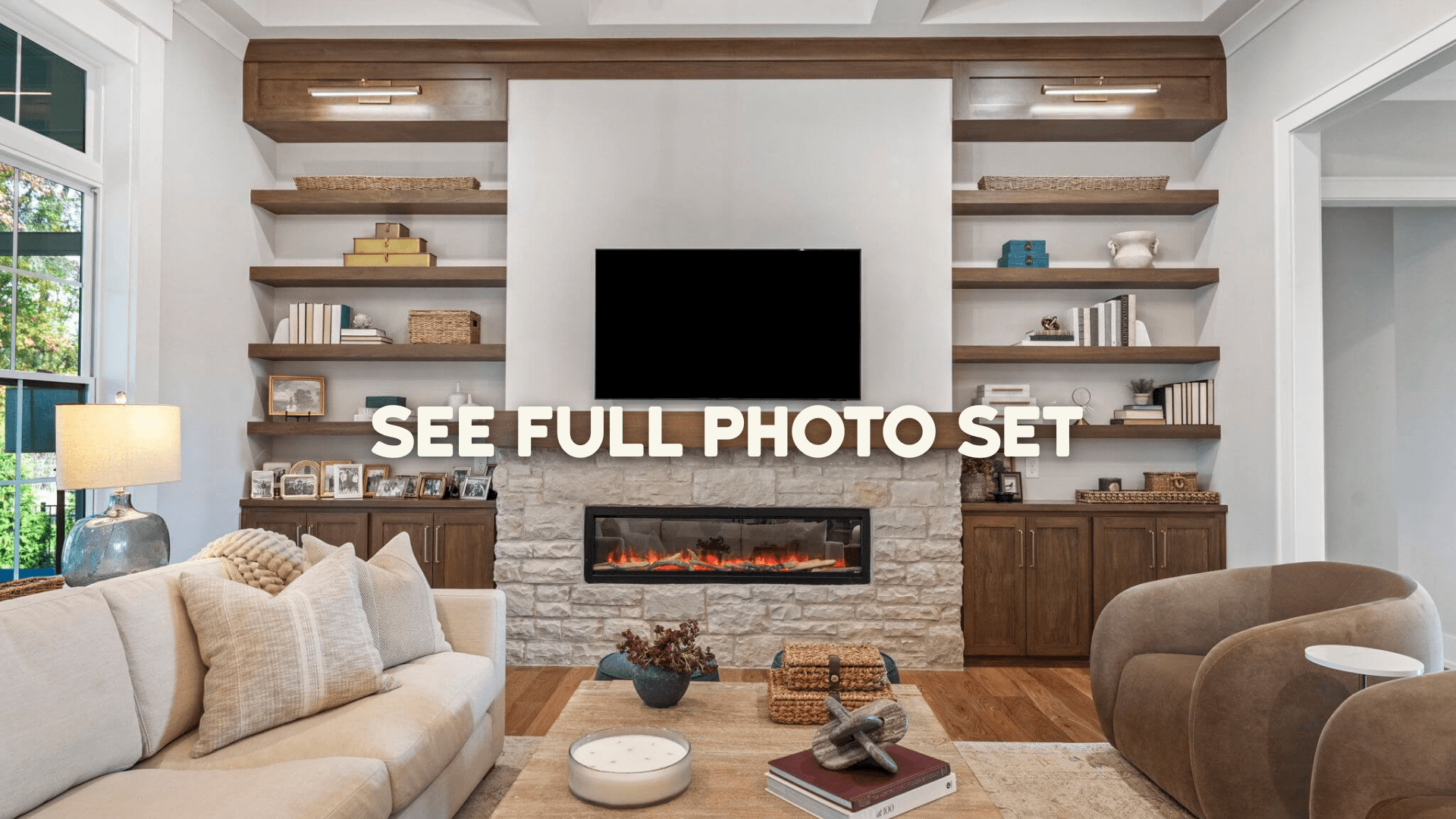 Modern living room with beige sofa, two armchairs, a stone fireplace with a mounted TV, and built-in wooden shelves with decorative items.
