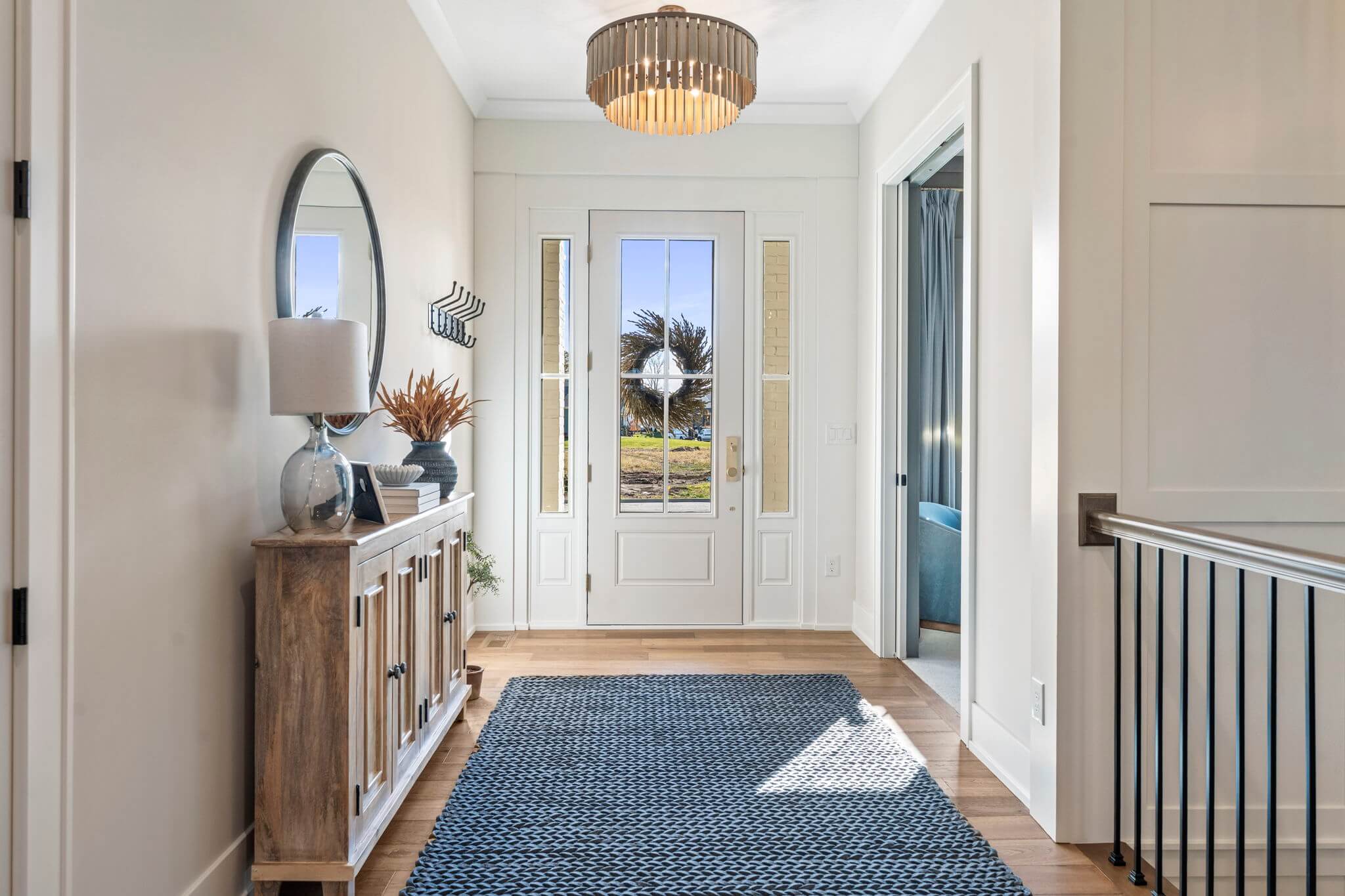 Bright entryway with wooden sideboard, round mirror, modern lamp, blue woven rug, and white front door with glass panels and wreath.