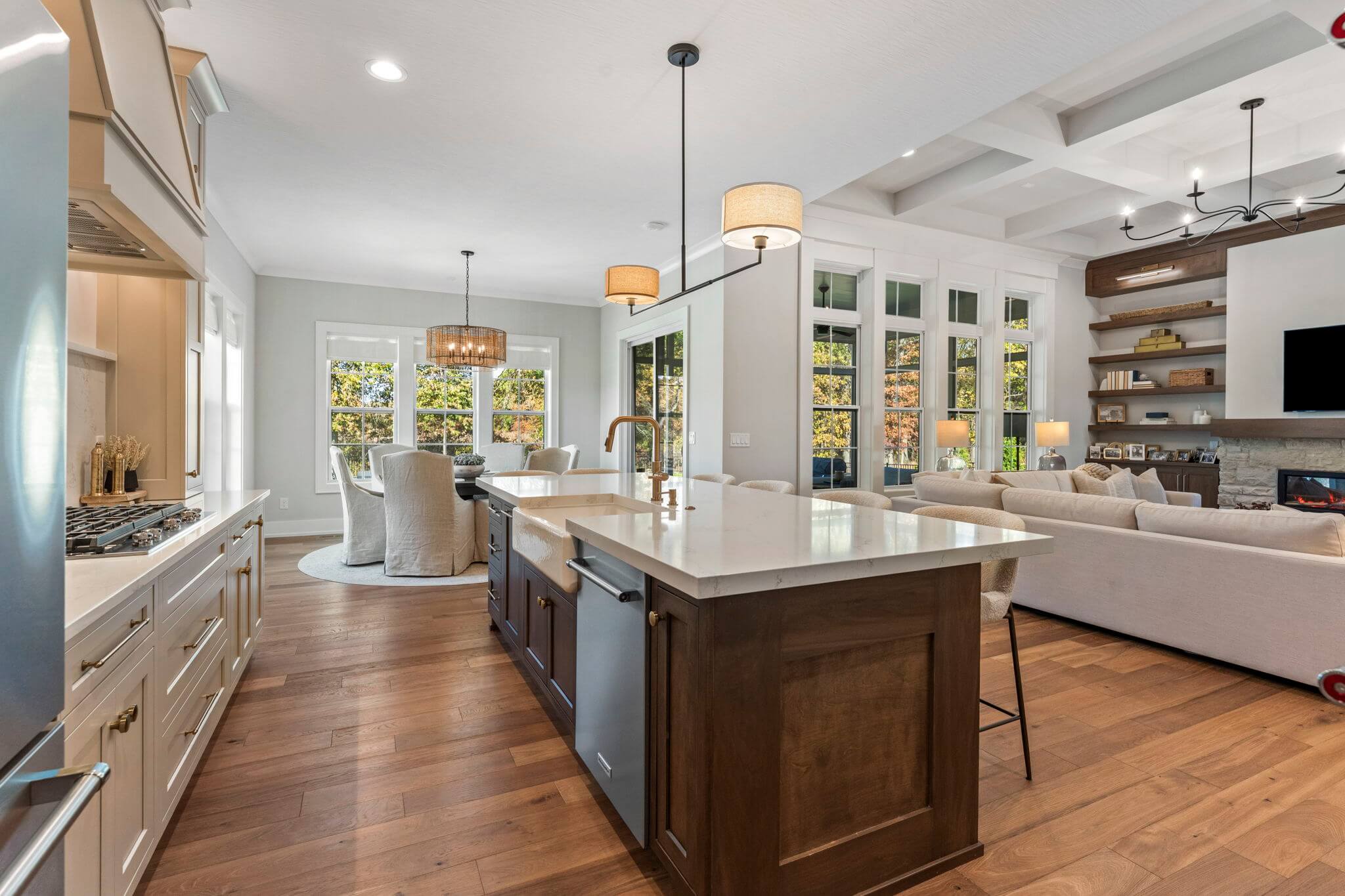 Open-concept kitchen and living room with a large island featuring a farmhouse sink, bar stools, hardwood floors, and large windows with views of autumn trees.
