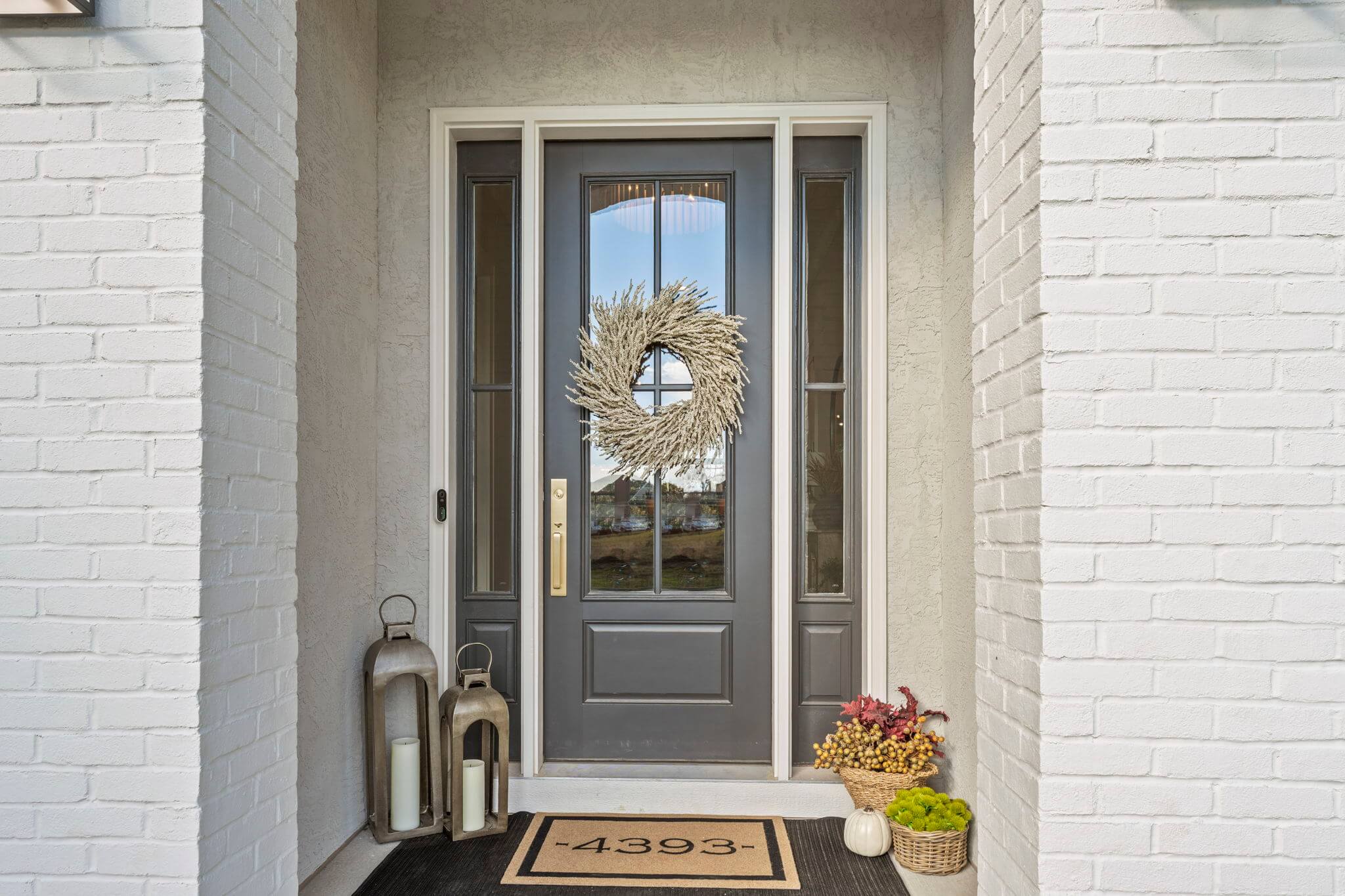 Gray front door with glass panels, decorated with a white wreath, flanked by two large lanterns with candles, a doormat with house number 4393, and baskets with fall foliage and greenery.