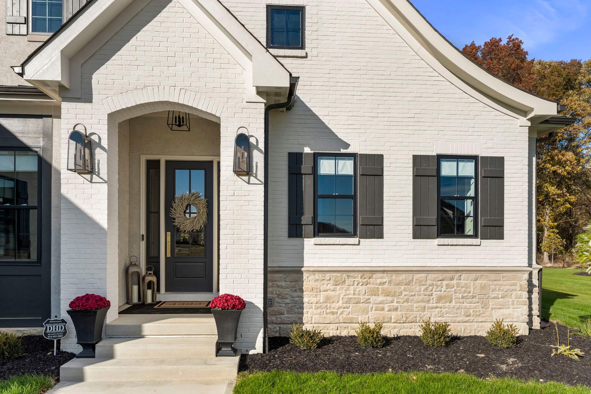 Front entrance of a white brick house with a black door adorned with a wreath, two side windows with black shutters, and red flowers in black planters on either side of the steps.
