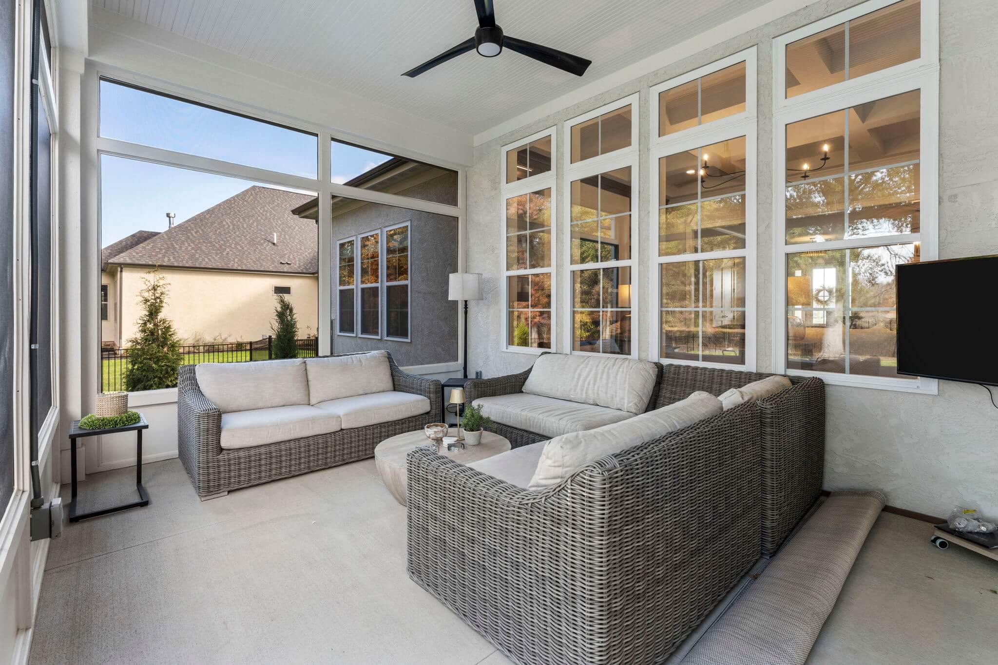 Screened-in porch with gray wicker sectional sofa, matching loveseat, round coffee table, ceiling fan, and large windows showing outdoor view.