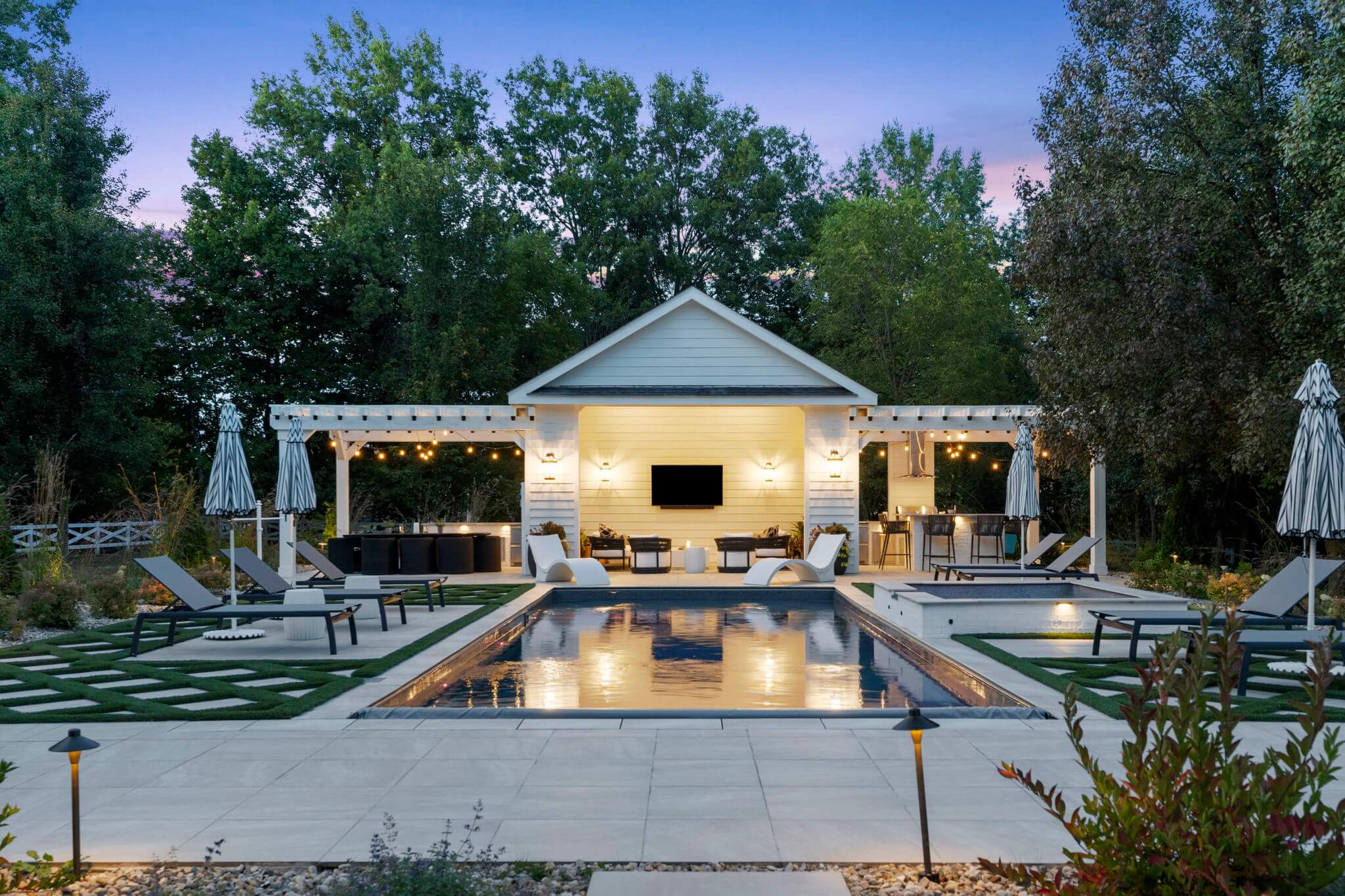 Evening view of a modern backyard with a lit swimming pool, white lounge chairs, striped umbrellas, and a pergola-covered seating area with a TV.