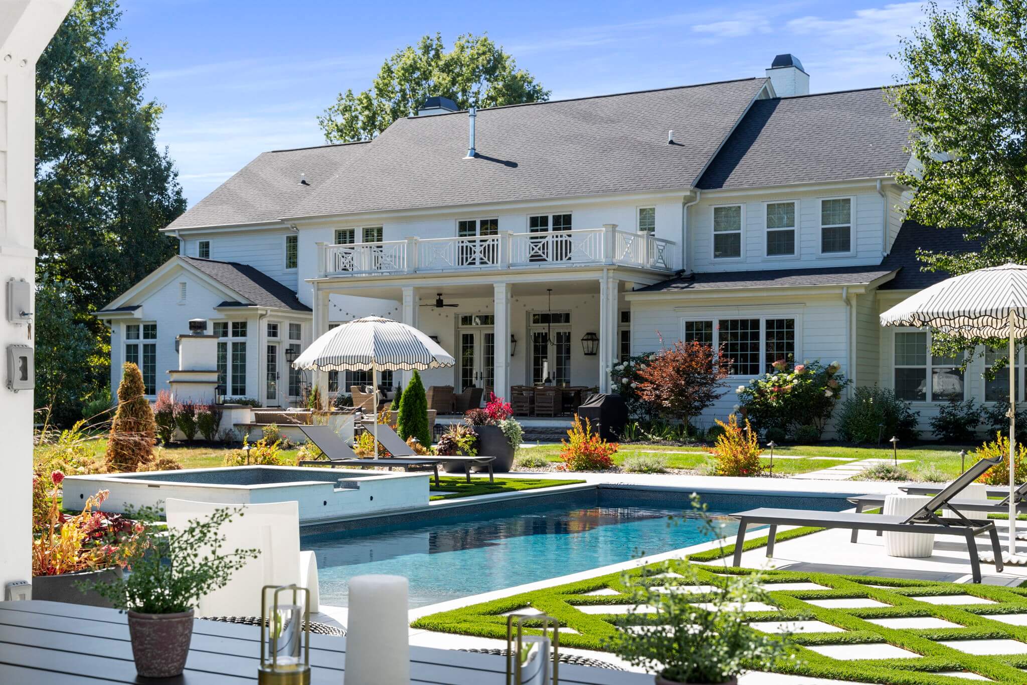 Large two-story white house with a gray roof, backyard pool, lounge chairs, striped umbrellas, and landscaped garden.