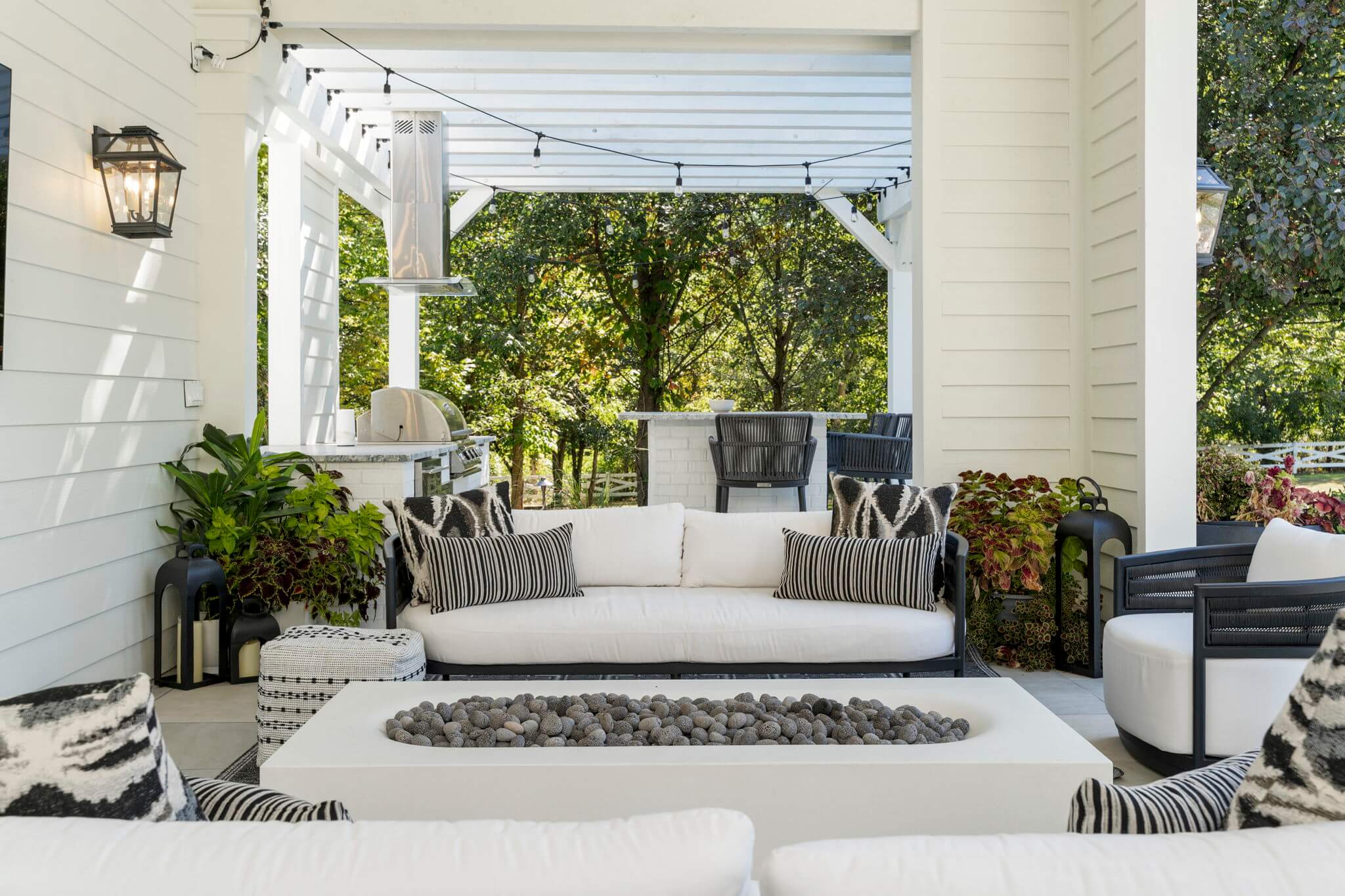 Modern outdoor patio with white cushioned sofas, black and white patterned pillows, a rectangular fire pit table filled with stones, and a pergola-covered kitchen area with bar stools surrounded by trees.