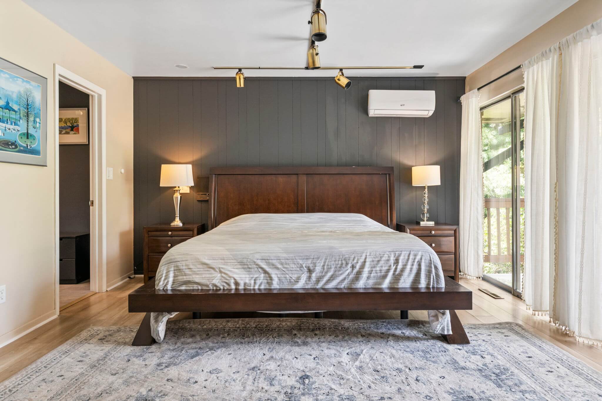 Modern bedroom with a dark wooden bed frame, two bedside tables with lamps, dark paneled accent wall, and sliding glass door with white curtains.