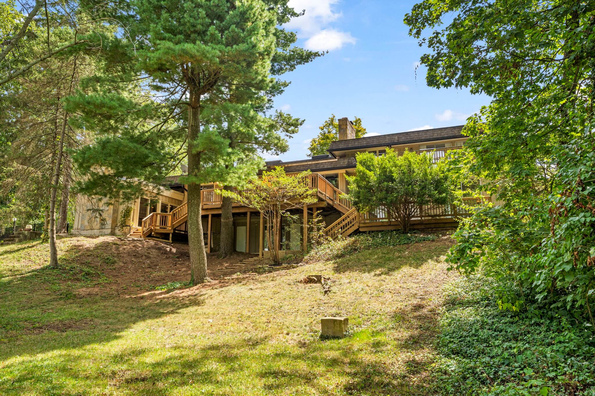 Backyard view of a house with an elevated wooden deck, surrounded by tall trees and greenery under a blue sky.