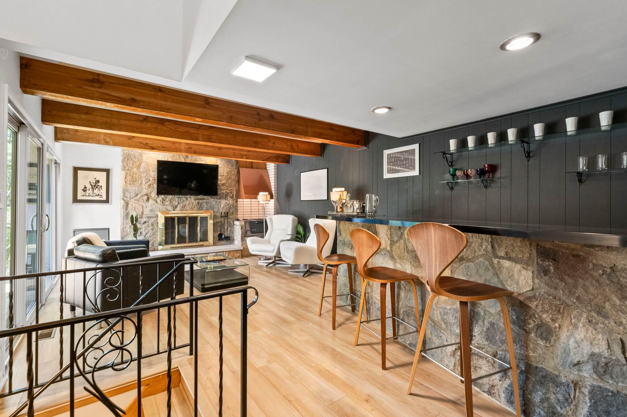 Modern living room with stone fireplace, wall-mounted TV, wooden ceiling beams, bar with wooden stools, and large glass sliding doors.