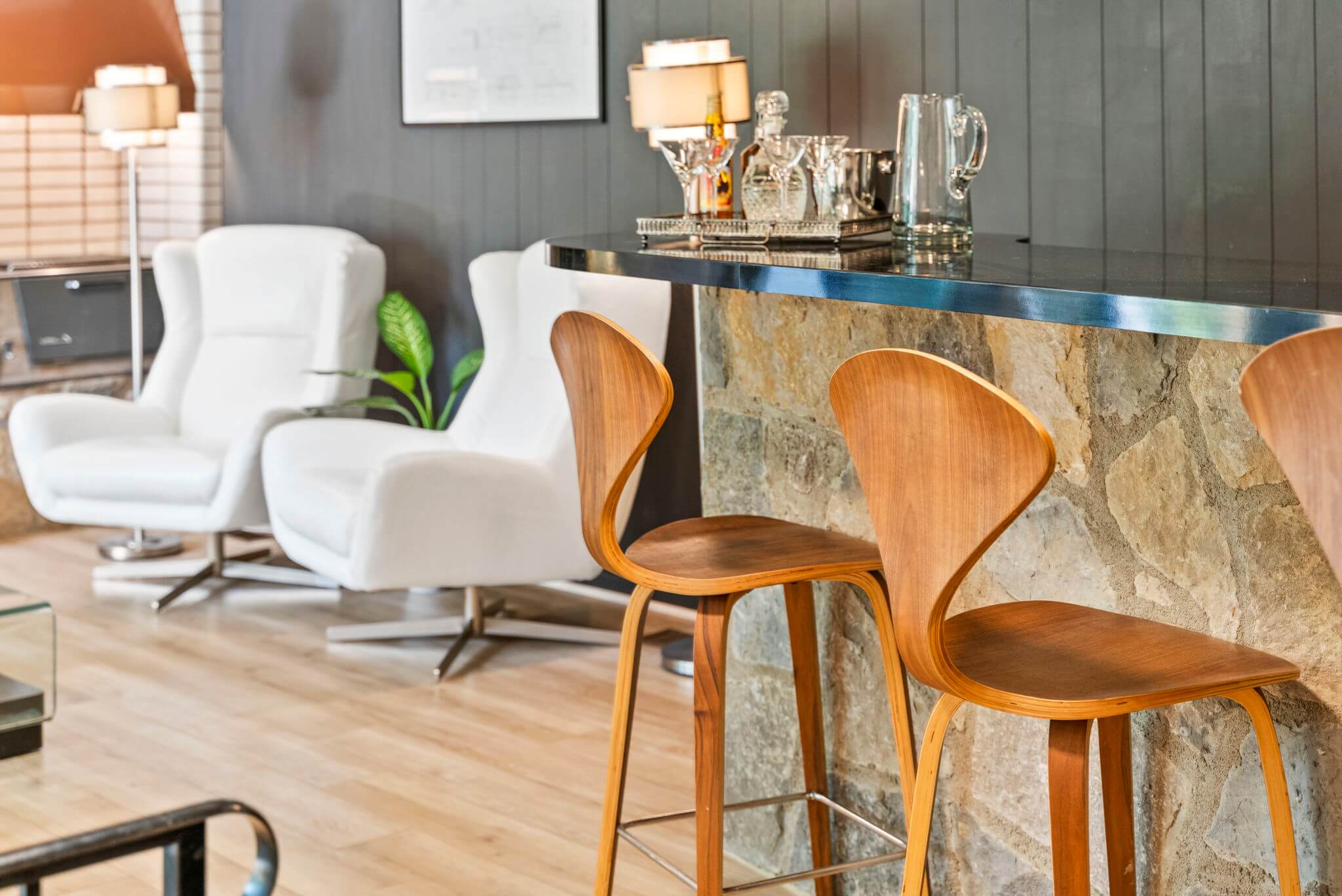 Modern bar area with wooden stools, stone countertop, and white armchairs in the background.