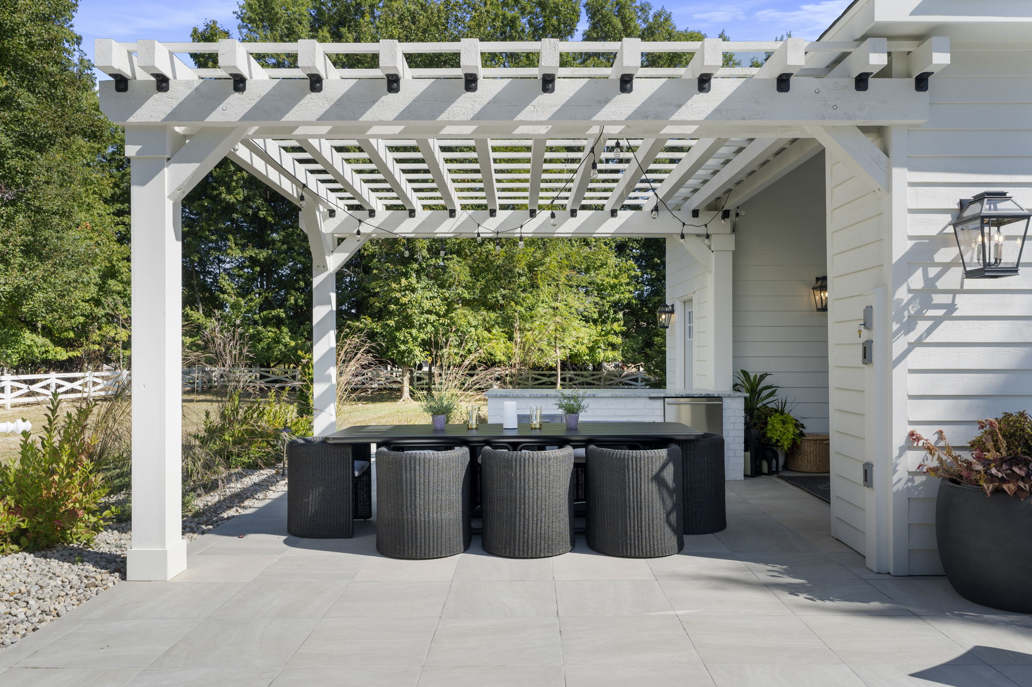 Outdoor dining area with black woven chairs and table under a white pergola on a tiled patio, surrounded by greenery.