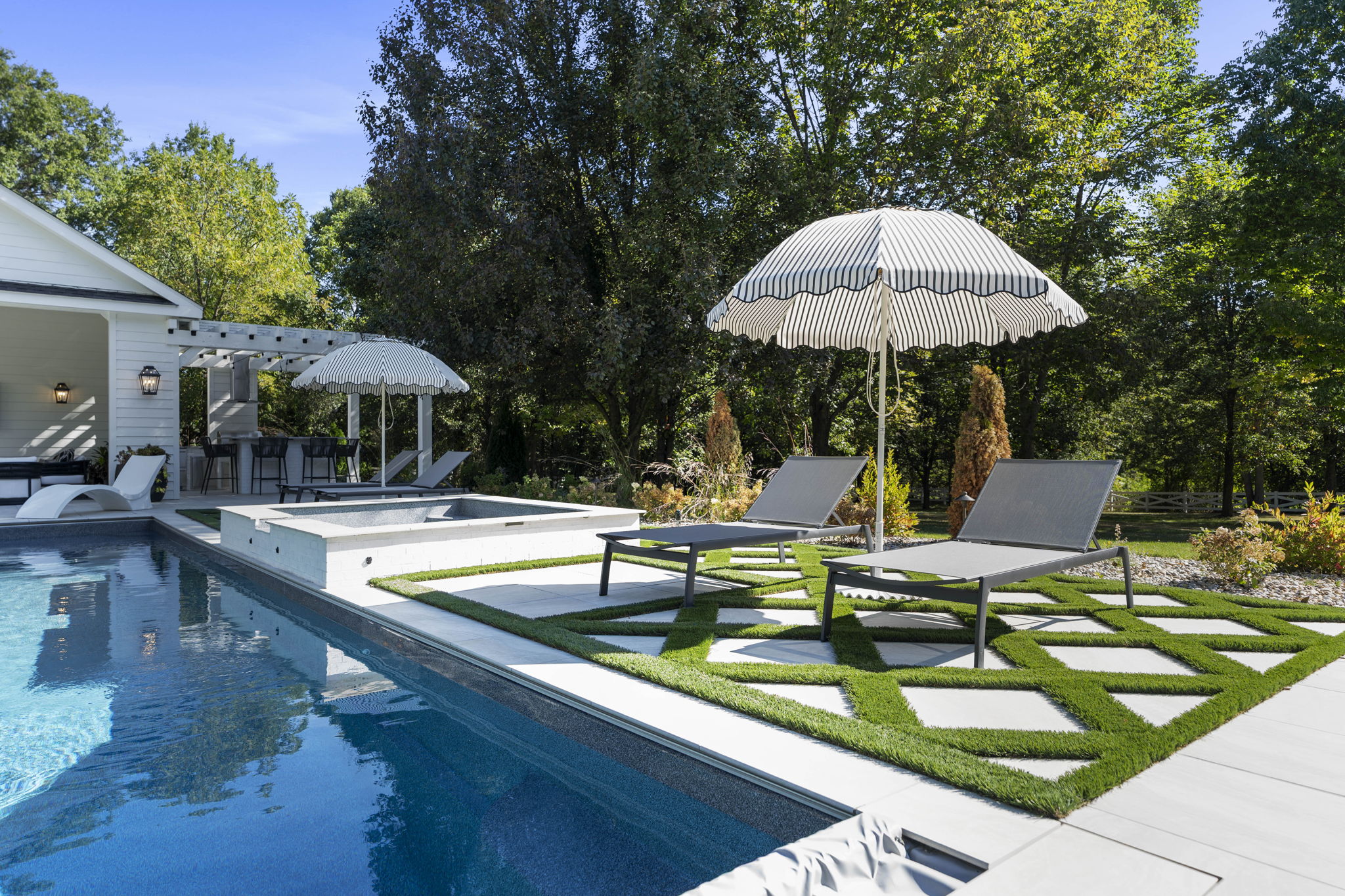 Modern backyard with a blue swimming pool, white hot tub, striped umbrellas, and gray lounge chairs on patterned grass and tiles.