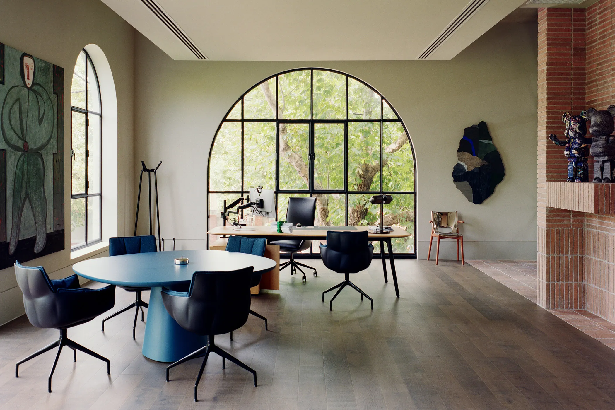 Modern office with a large arched window, a blue round table with black chairs, a wooden desk with two office chairs, and wall art.