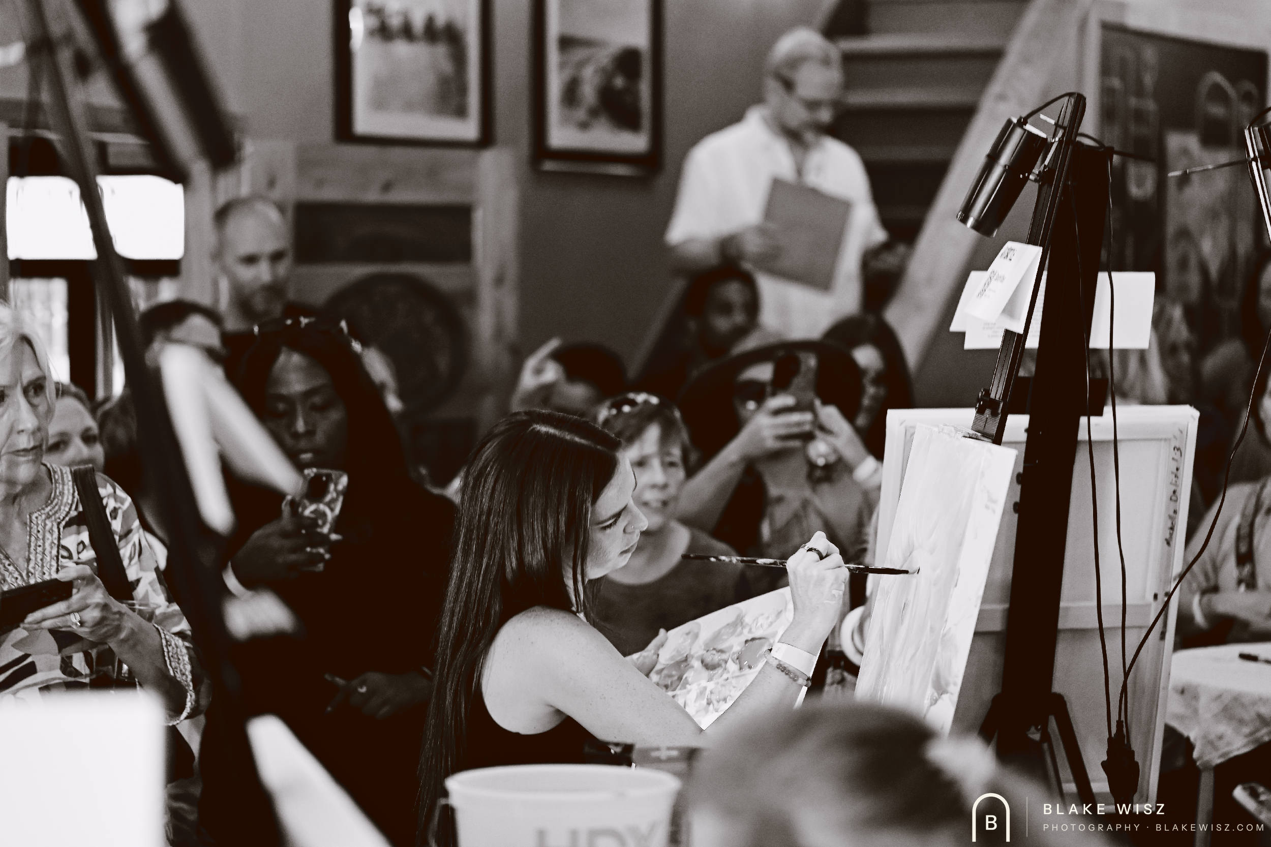 Black and white photo of an artist painting while the crowd photographs her round at Ormond Brewing
