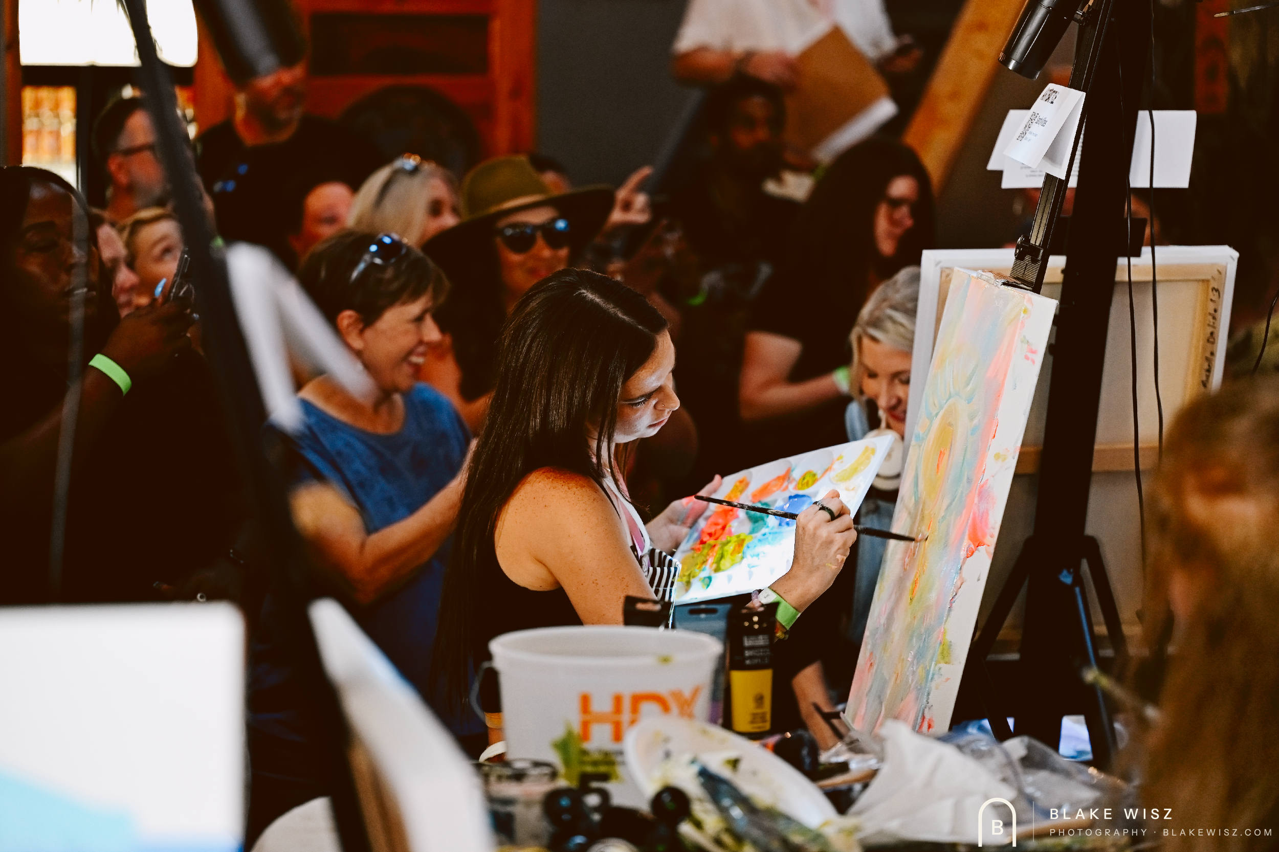Artist holding a loaded palette while painting a pastel canvas as spectators watch from inches away