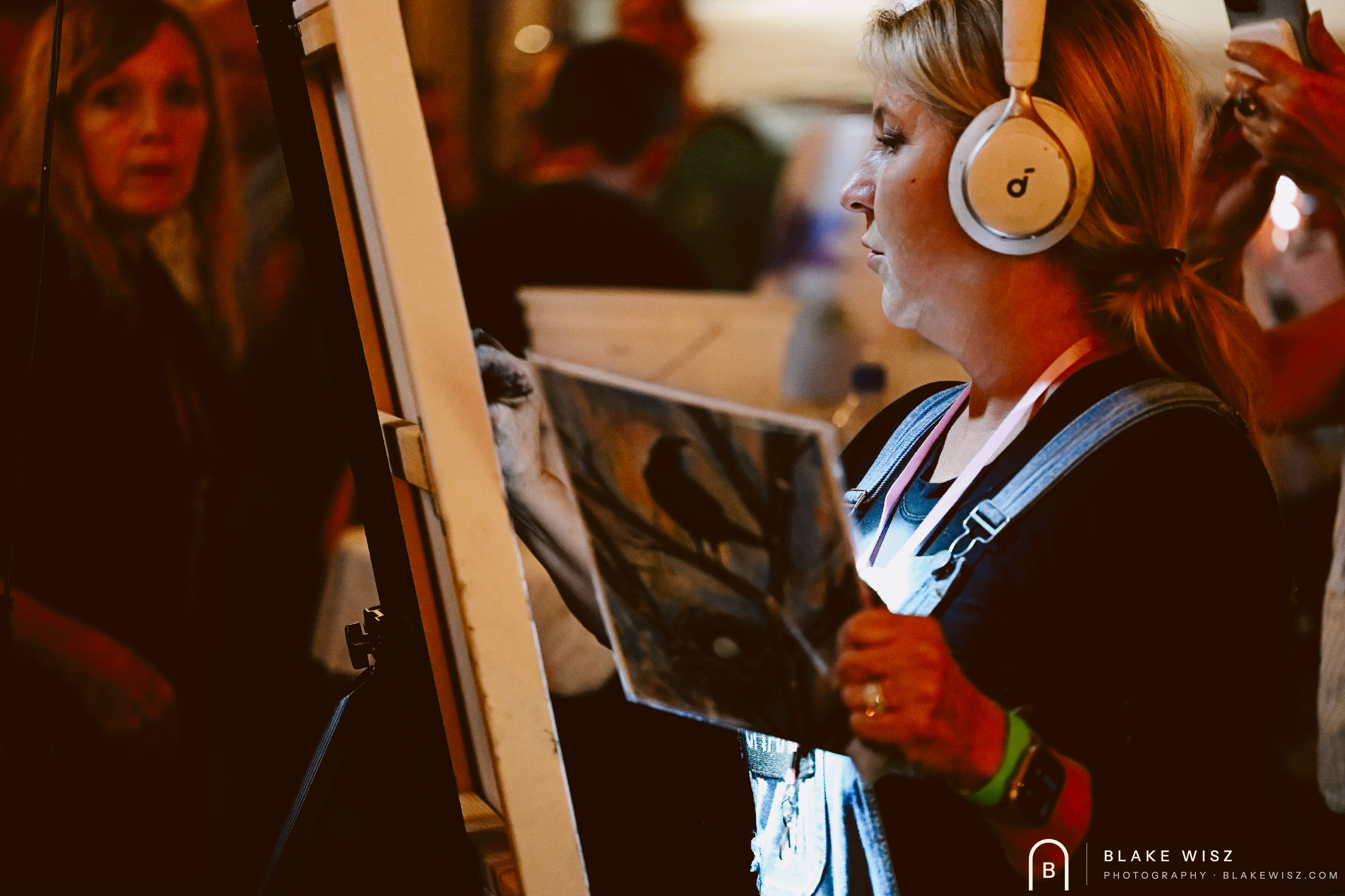 Michelle A Delisle at her canvas holding the bird reference print under warm venue lighting
