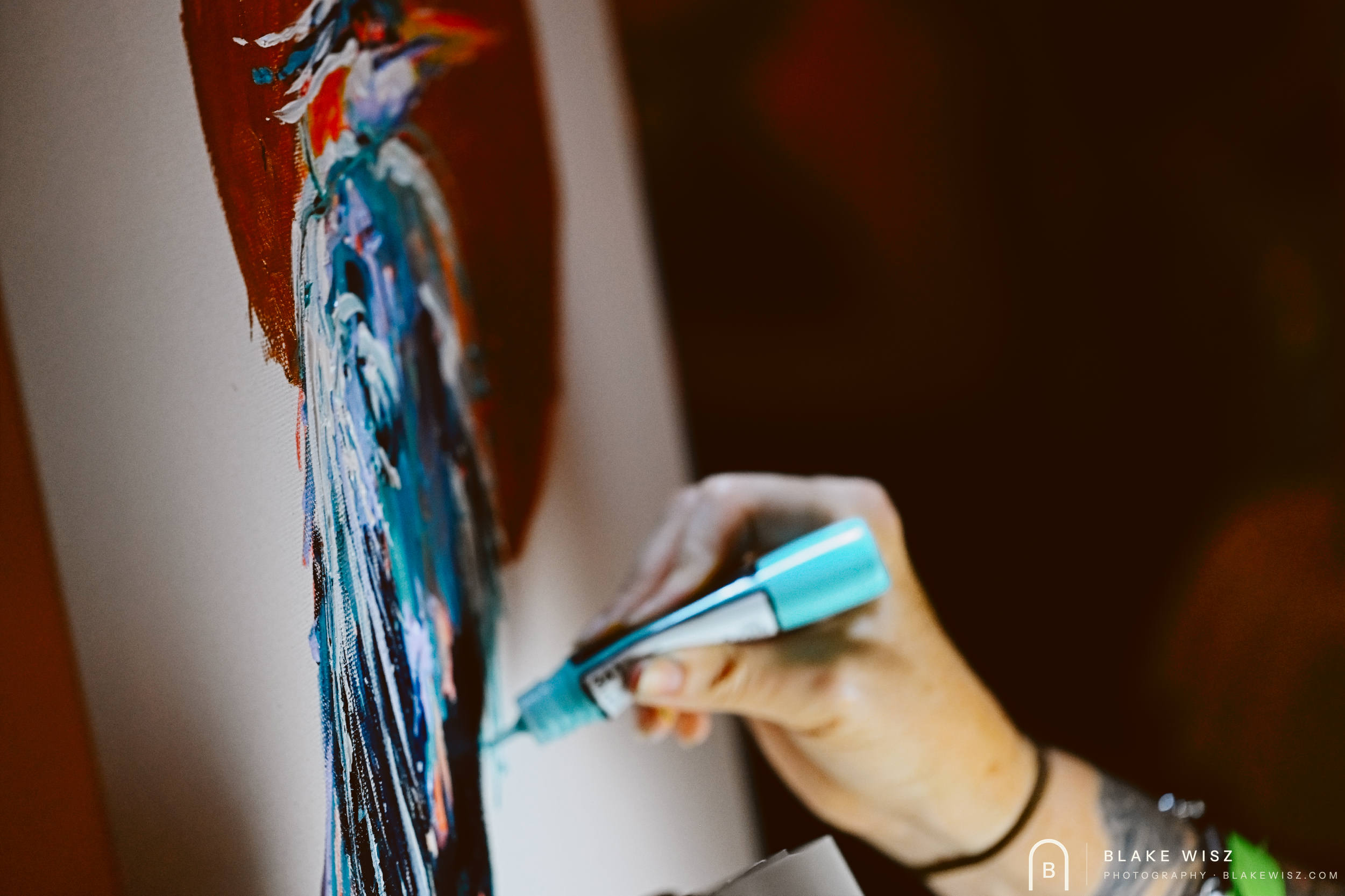 Artist applying teal paint from a marker to a heron painting on a deep red ground